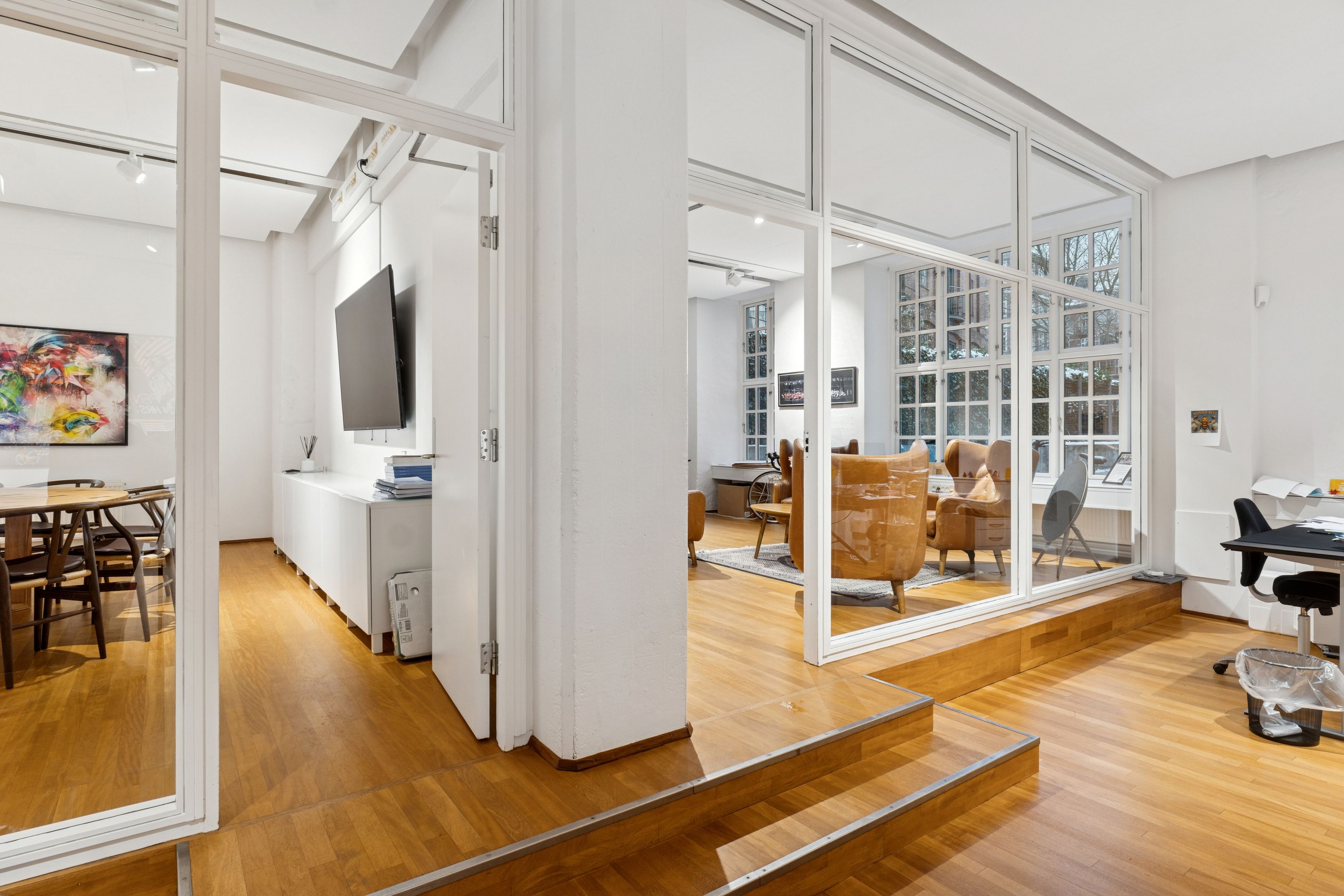 Vider åbning med glasvinduer og højloft til kontorlokaler med lysegulve og møbler i lyse nuancer samt udsigt til gaden