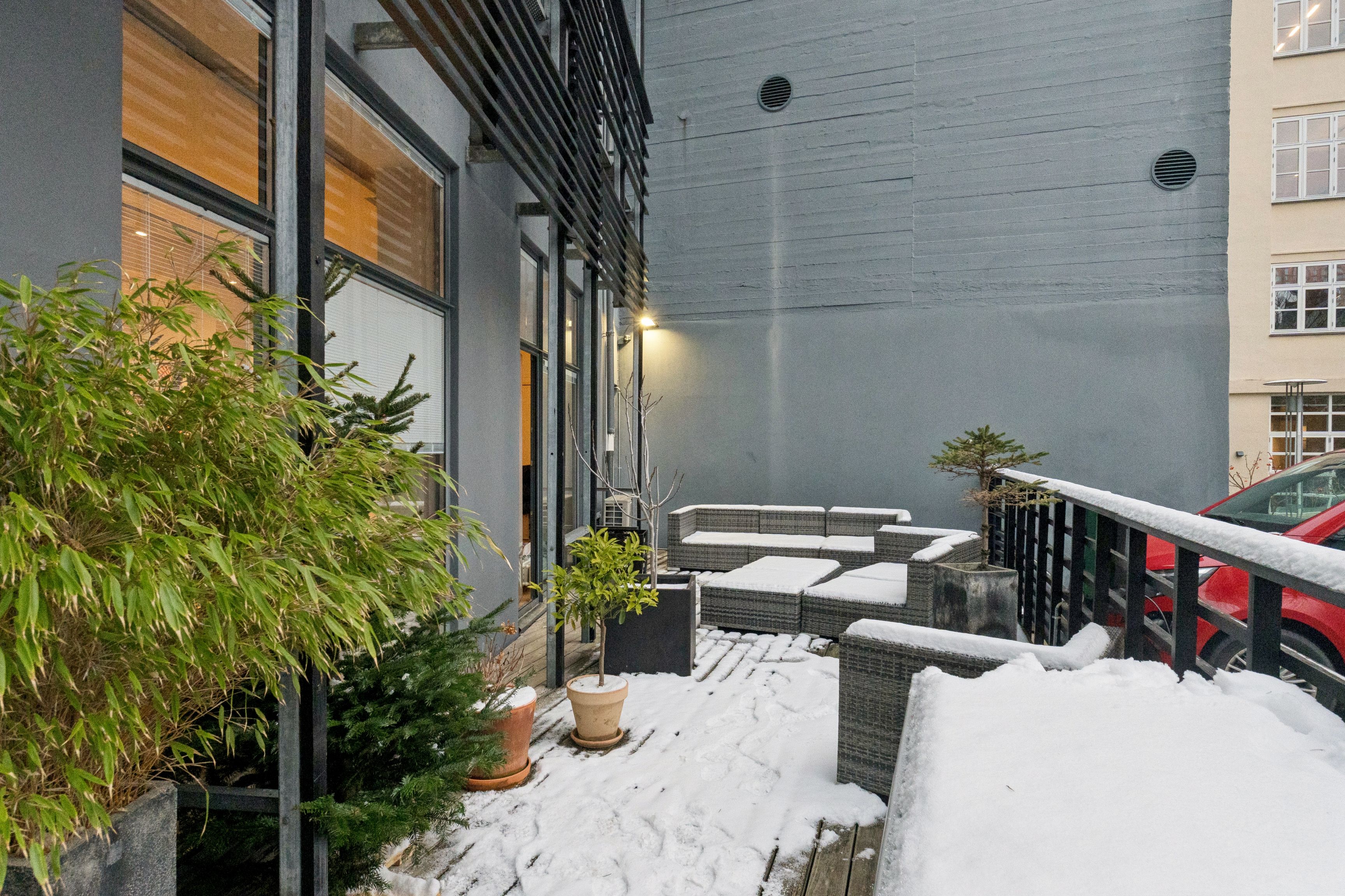 Privat terrasse med snedækket og plantepot med sofa og stol samt redskaber og sne samt et bygningsvæg med vinduer og indgang