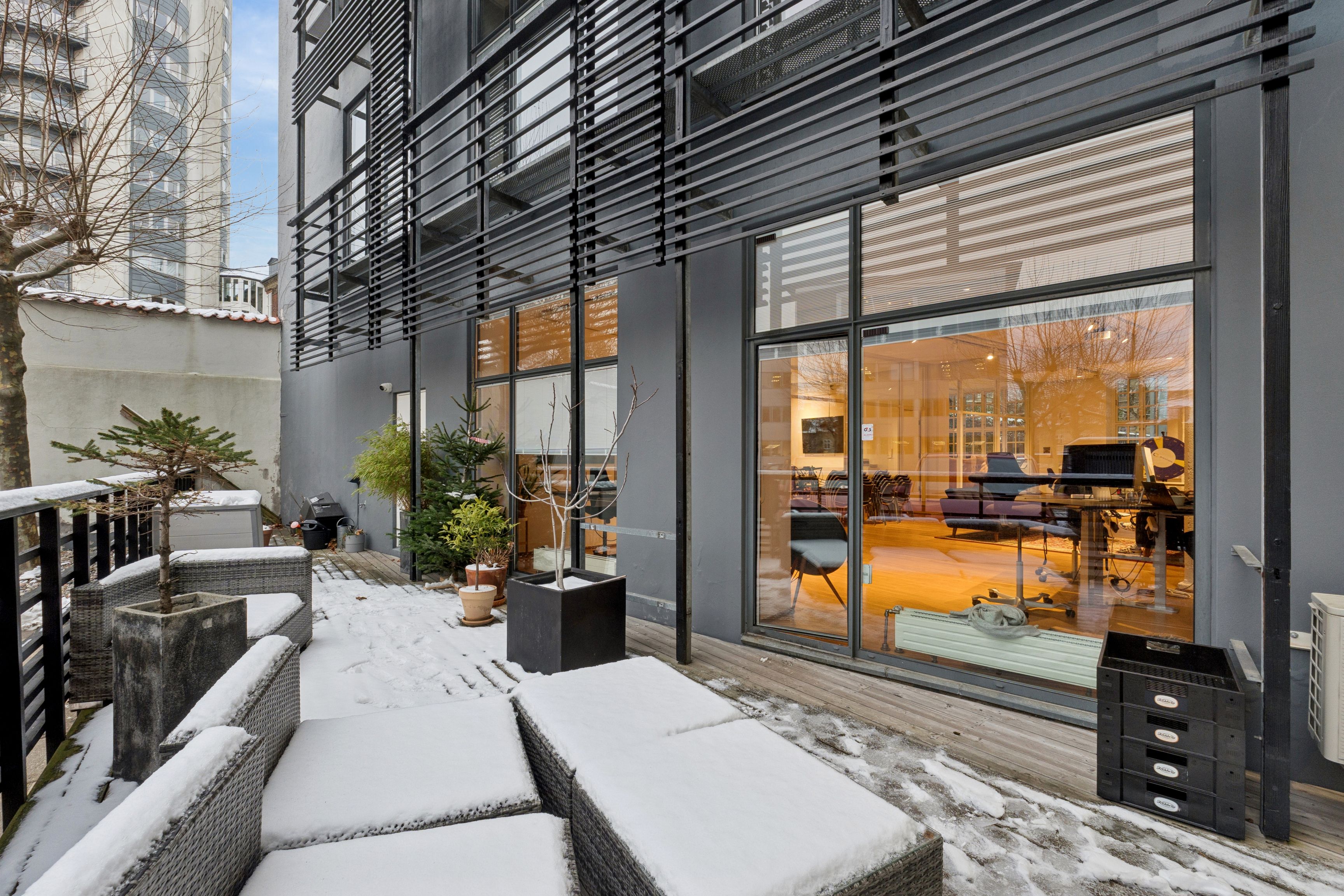 Moderne kontor med høj loftshøjde og privat terrasse med snedækket og plantepoteter, udsigt til en gul gulv og møbler samt udsigt til en høj bygning med store vinduer