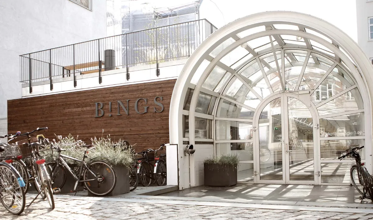 En række cykler står parkeret ved et moderne kontorbygning med en lys, buet indgang på Vesterbrogade 149. B!NGS står skrevet i store bogstaver på facaden