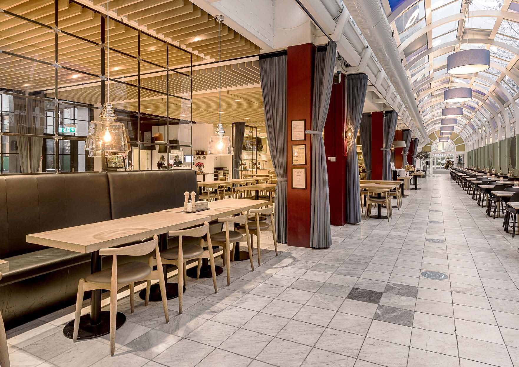 Lyst og moderne kontor med høj loftshøjde og privat terrasse. Væggene med glas og lysekroner. Bord og stole er stillet op til møder. Lamper hængende fra loftet.