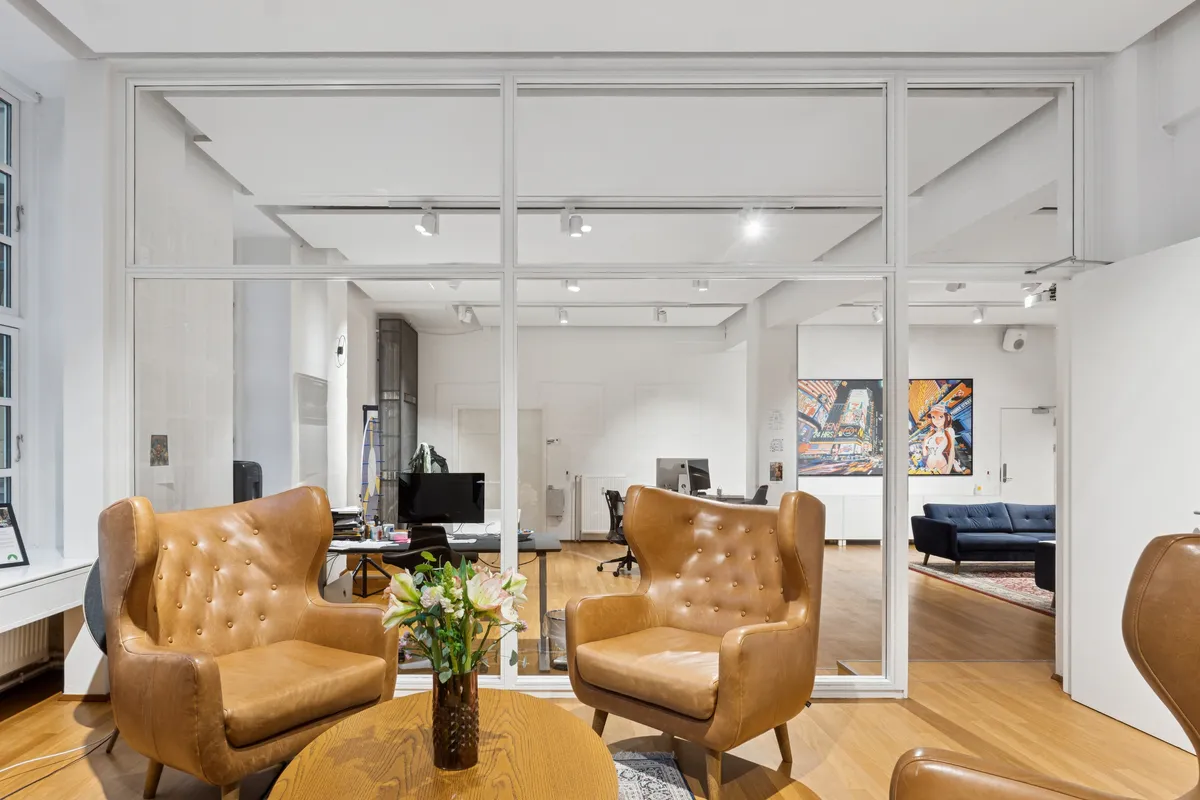 Lyst og moderne kontor med høj loftshøjde og privat terrasse. Kontoret har to læderstole og et gult rundt bord med blomster.