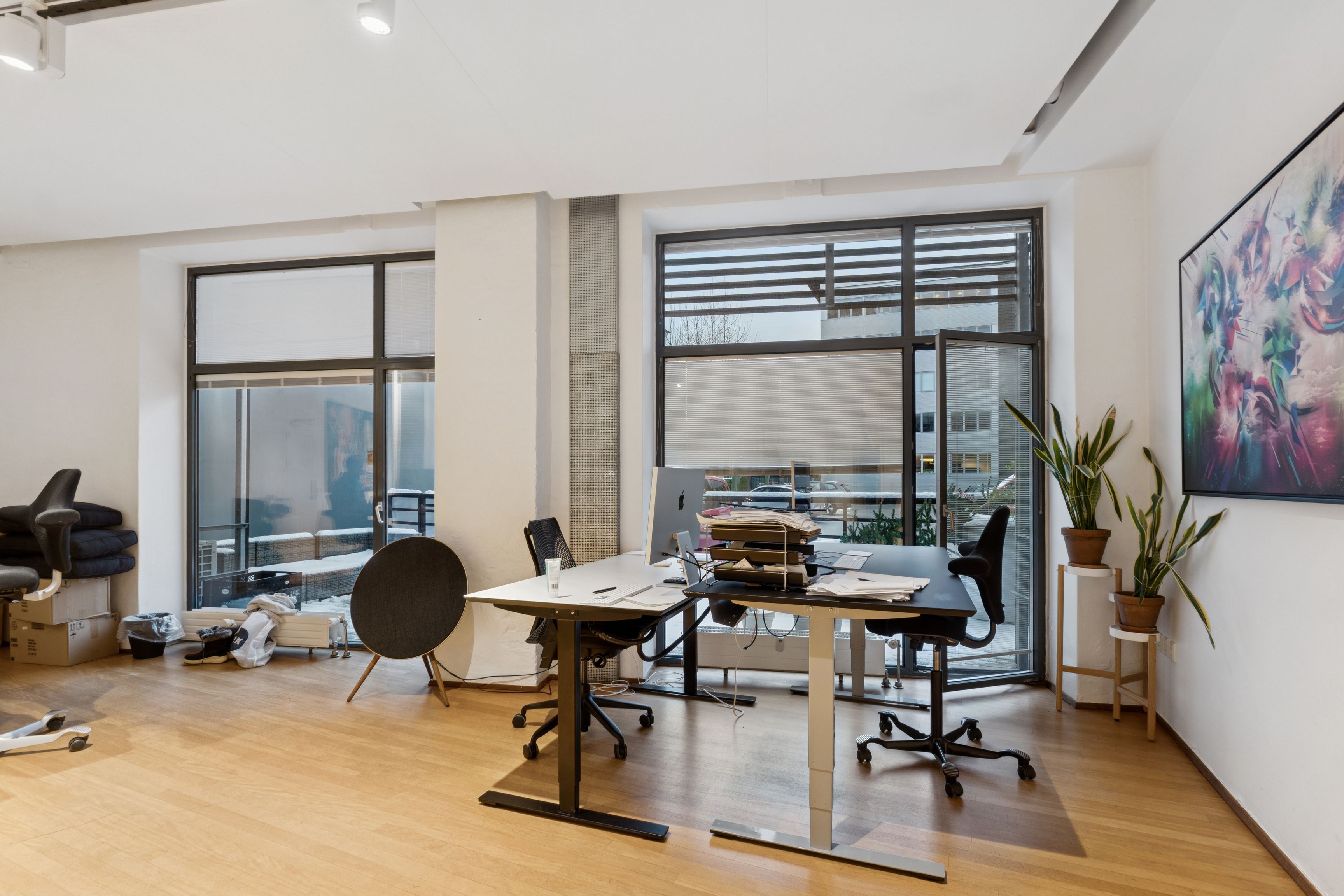 Lyst og moderne kontor med høj loftshøjde, skriveborde, kontormøbler, terrasse, vinduer og planter mod en stor bygning med snedækkede områder i baggrunden