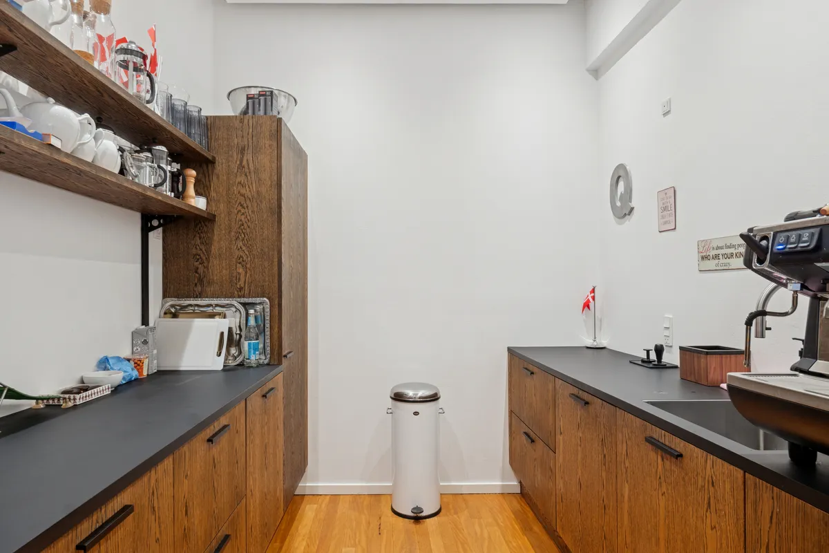 Lyst og moderne kontor med høj loftshøjde og privat terrasse. Køkken med træskabe, sort bordplade og støjle. Moderne kaffemaskine og hvid skraldespand. Loftet er højt og lyse.