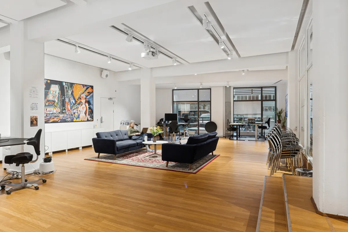 Moderne kontorlokaler med høj loftshøjde og lyse rum, der indeholder skriveborde, møbler og terrasse med udsigt til gaden