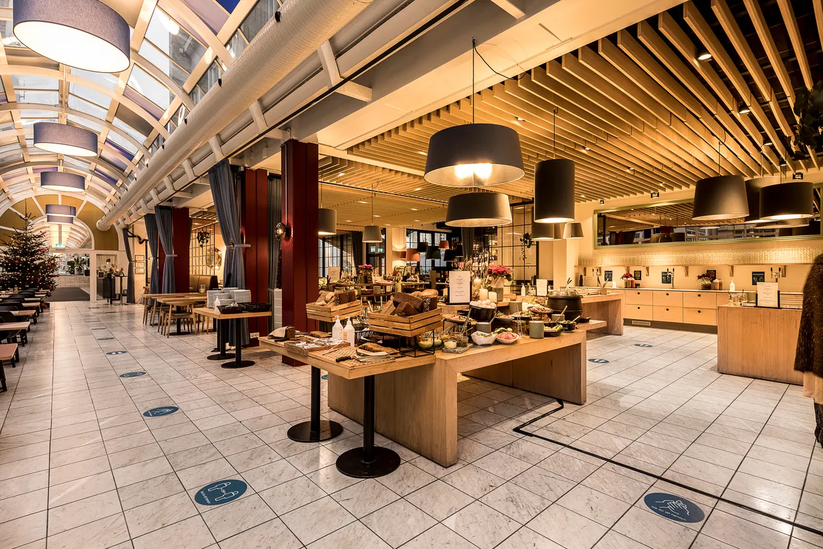 Moderne kontorloft med loftshøjde, lyse søjler og lysekroner, serveringsbord med retter og drikke, hvid flisegulv, kryds og pil for social afstand, glasvinduer med udsigt og juletræ