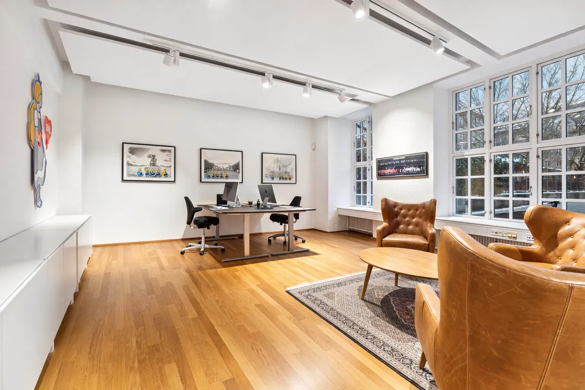 Lyst kontor med højloft, moderne skriveborde og tre lejlighedsmøblerede stole i læder samt to store vinduer med udsigt til gaden og træer