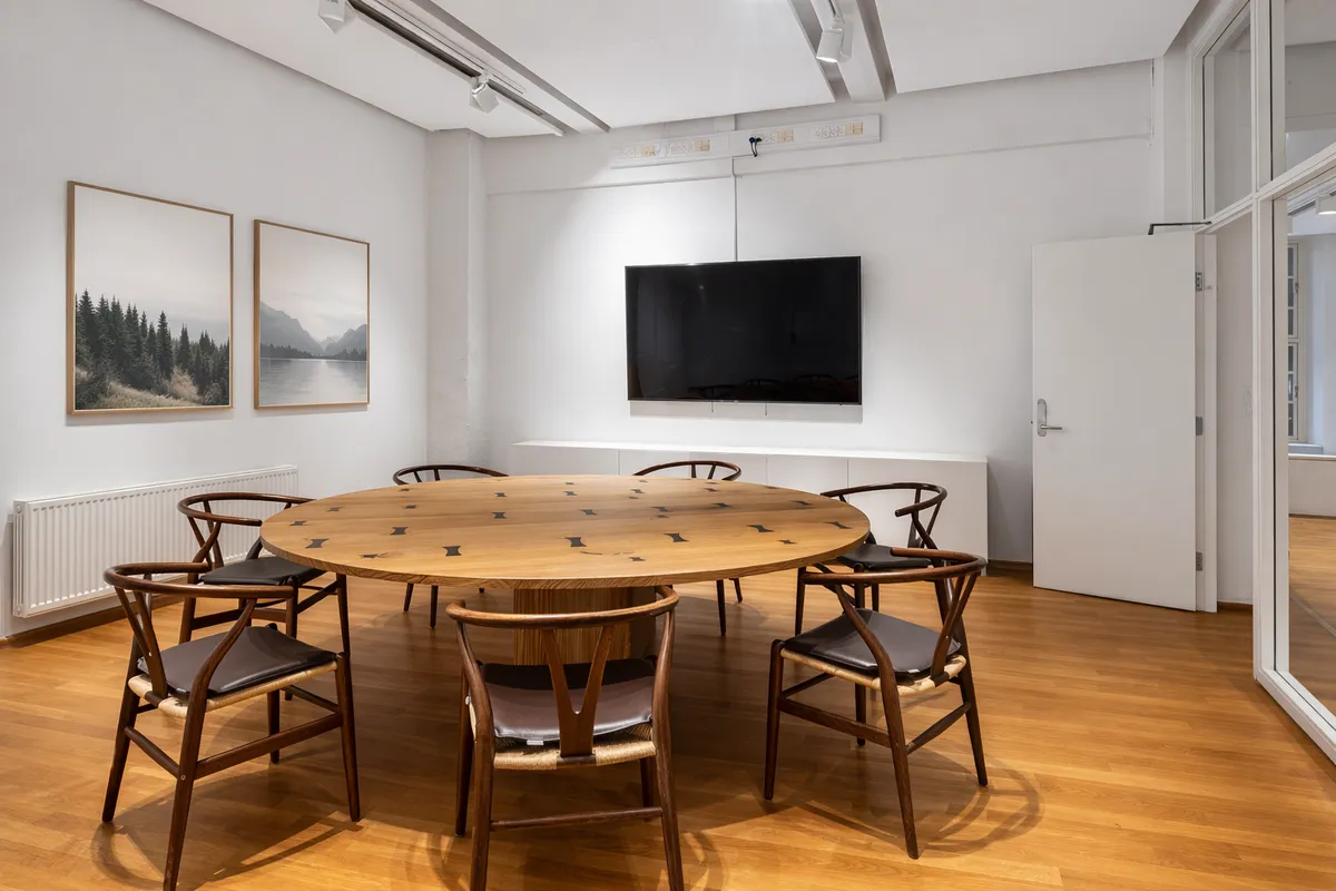 Rundt bord med otte stole omkring, indretning med tv-skærm, hvide vægge, to billeder med landskaber og skov, hvide gardiner, høj loftshøjde og åben dør til terrasse