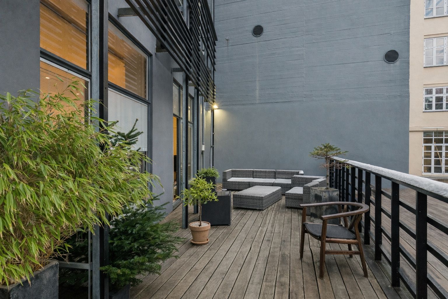 Terrasse med moderne møbler og planter. Sort metalgelænder og en lysekron. Bygning med vinduer og døre. Solskin og grønne planter på terrassen.