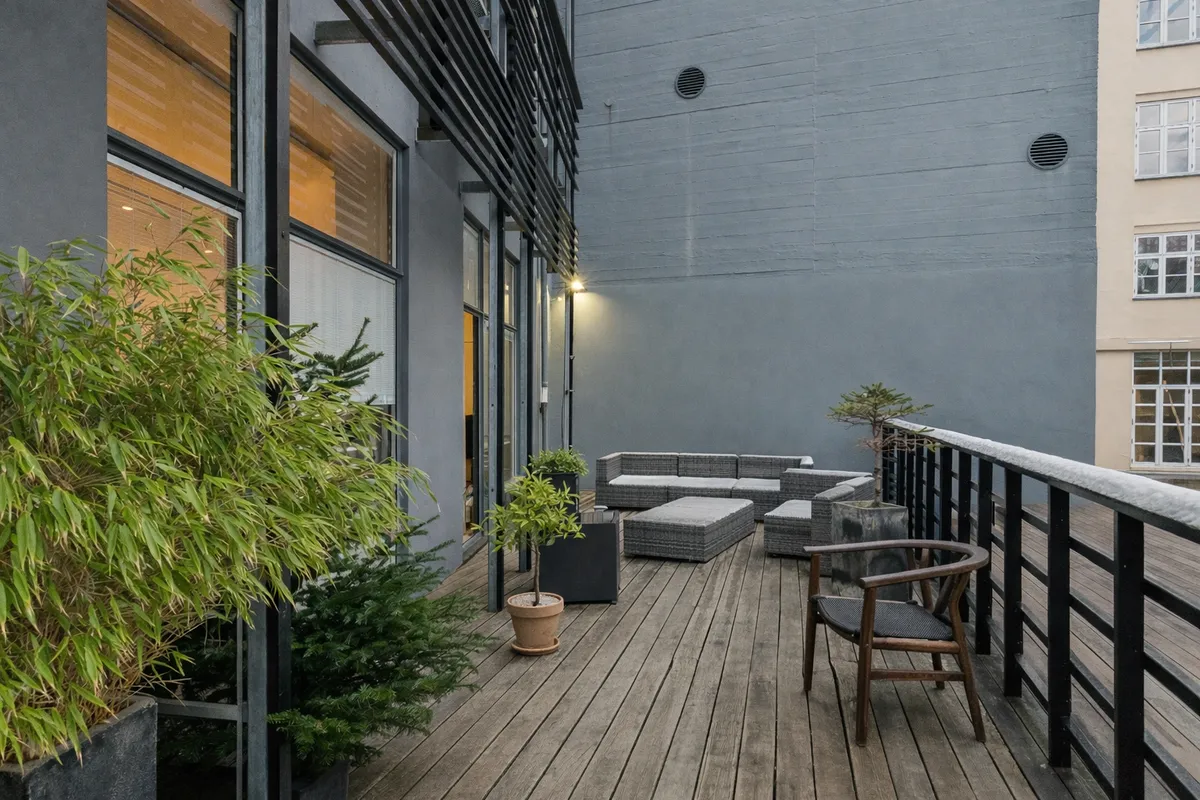 Terrasse med moderne møbler og planter. Sort metalgelænder og en lysekron. Bygning med vinduer og døre. Solskin og grønne planter på terrassen.