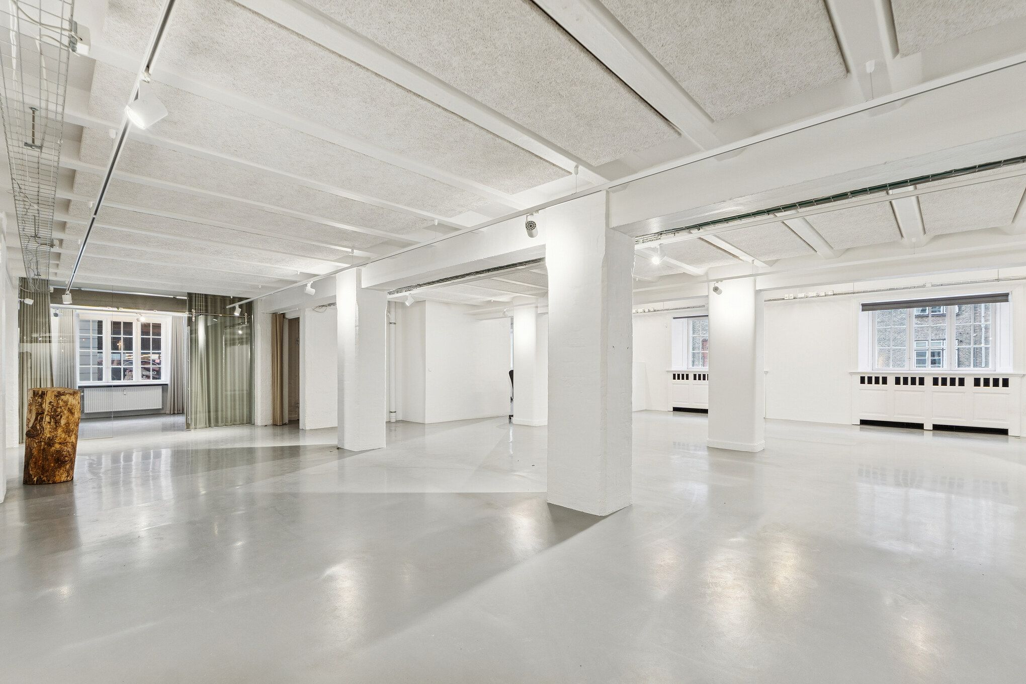 Moderne og industrielt kontor med hvide vægge og lysekroner, lysegulve og store vinduer mod gaden, samt træstamme og radiatorer i hvide vægge