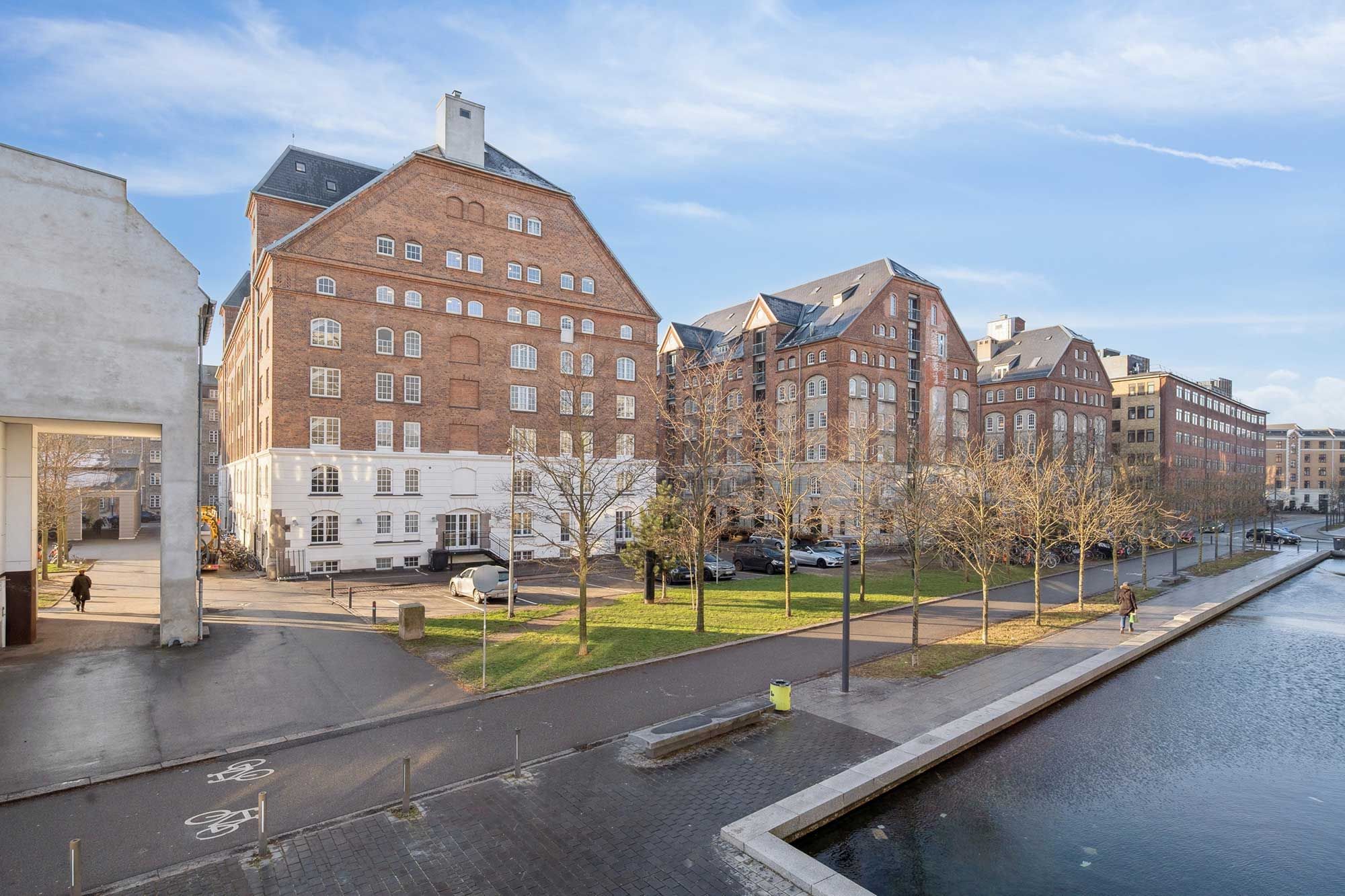 Kontor i Njalsgade med røde mursten, store vinduer og kanal langs siden. Parkerede biler og gående personer på gågaden. Bygninger med forskellige stilarter og facader.