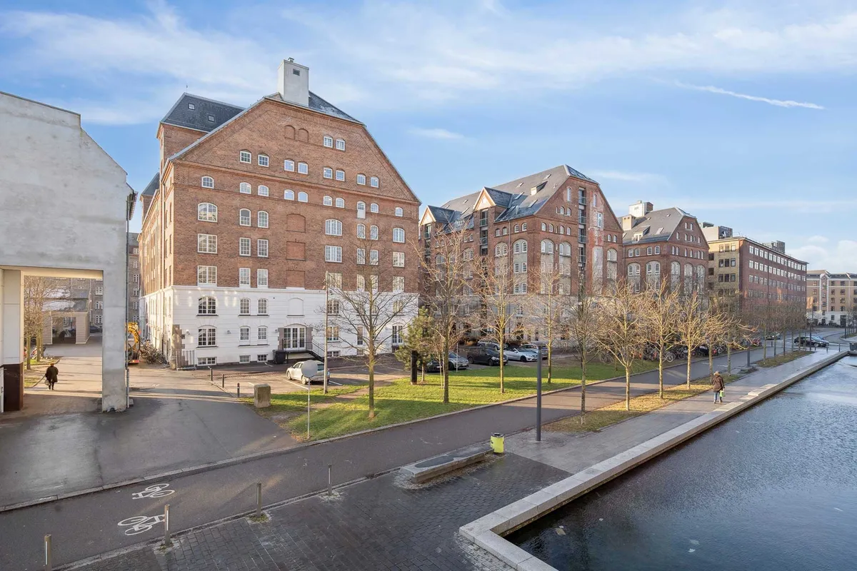 Kontor i Njalsgade med røde mursten, store vinduer og kanal langs siden. Parkerede biler og gående personer på gågaden. Bygninger med forskellige stilarter og facader.