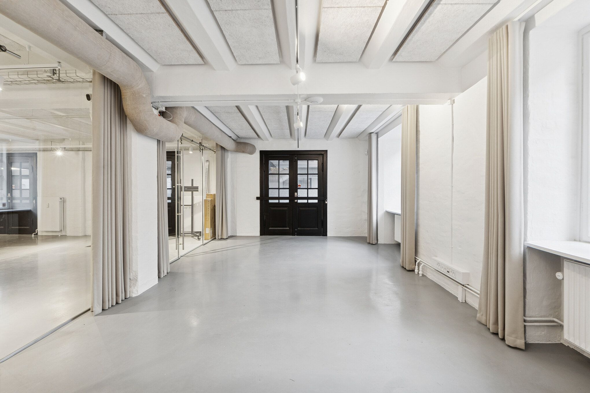 Minimalistisk kontor med hvide vægge, glasdøre, lysekroner, vinduer og møbler i lyse nuancer og hvid cementgulv samt højloftede rum med industrielle elementer