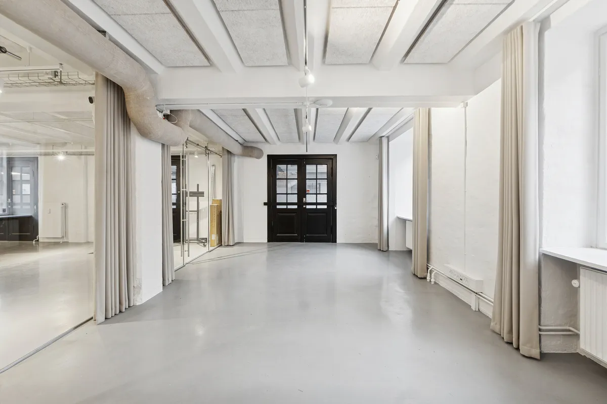 Minimalistisk kontor med hvide vægge, glasdøre, lysekroner, vinduer og møbler i lyse nuancer og hvid cementgulv samt højloftede rum med industrielle elementer