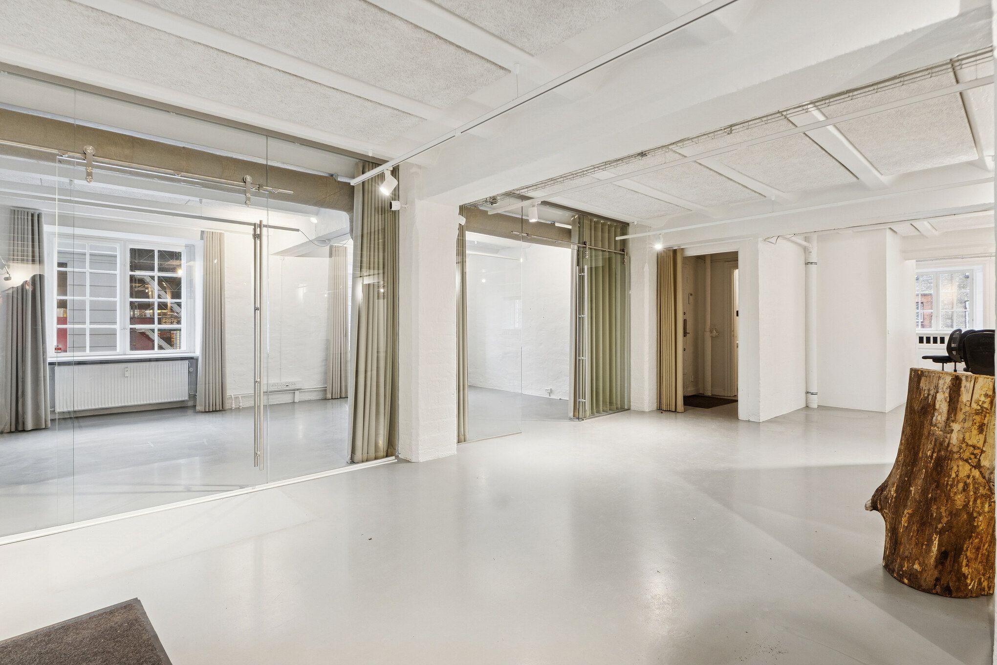 Rå og industrielt kontor med hvid gulv og vægge, store glasruder, åbne døre, moderne lampe, træstamme og gardiner, hvor glasruderne reflekterer rummet