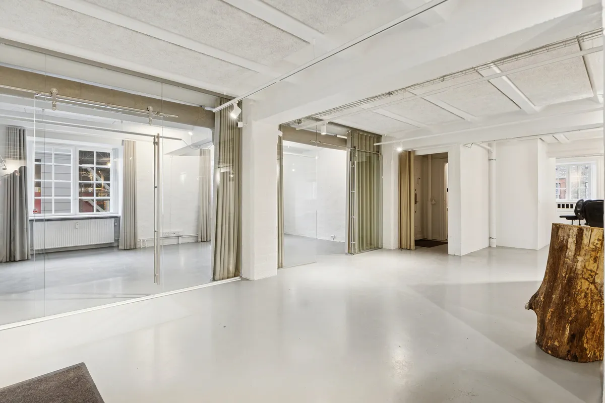 Rå og industrielt kontor med hvid gulv og vægge, store glasruder, åbne døre, moderne lampe, træstamme og gardiner, hvor glasruderne reflekterer rummet