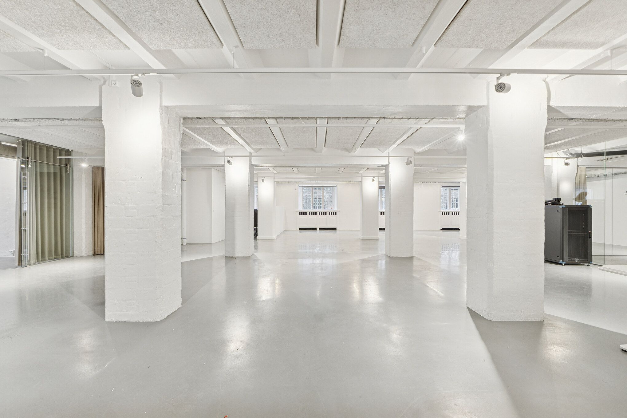 En stor, rummelig og hvid stue med lyse lofter, søjler og ren gulvoverflade samt moderne lysinstallationer. Et stort glasvindue og slanke hvide søjler giver et industrielt og minimalistisk udseende.