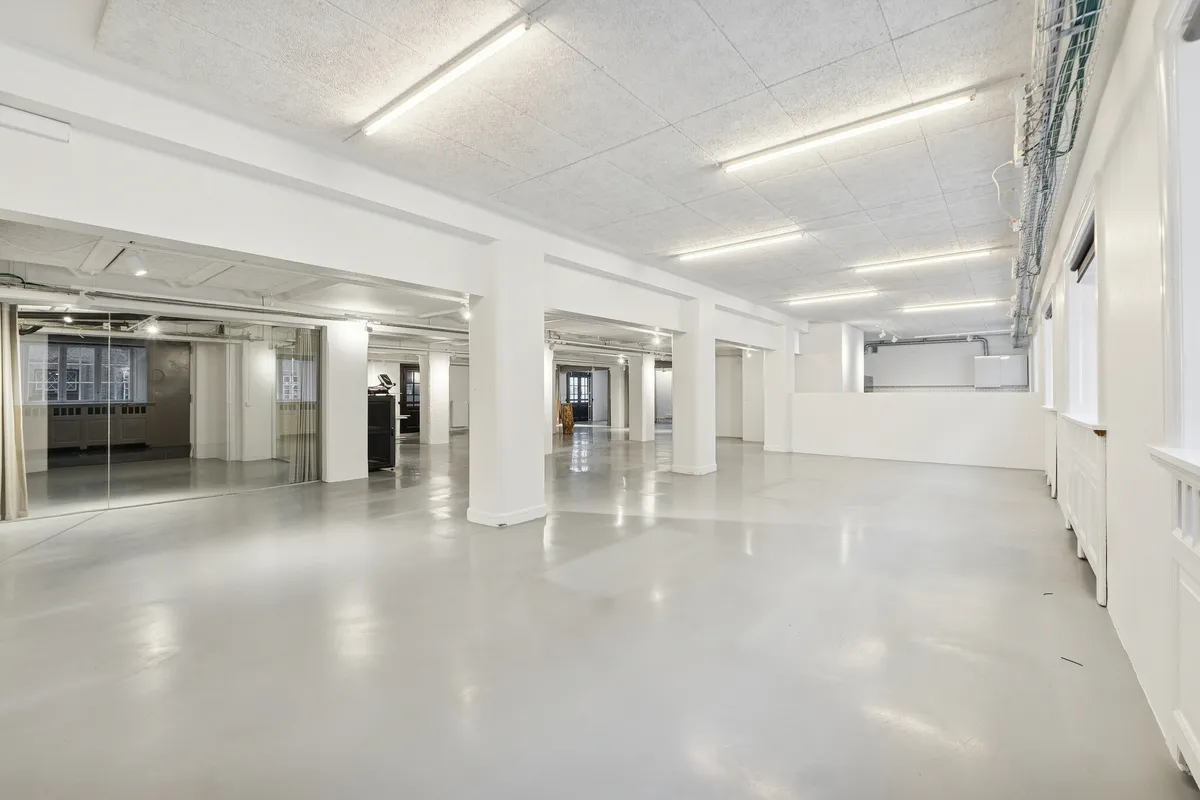 En stor, rå og industriel kontorrum med hvide vægge og loft. Der er en række lysekroner og hvidt marmorgulv. Der er en stor spejlramme og en dør i baggrunden