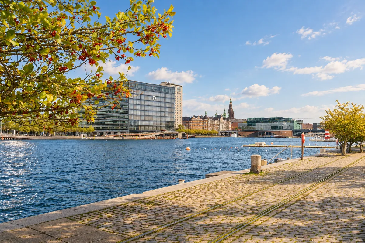 Københavns Islands Brygge med moderne bygninger og grønne træer langs vandkanten. Flod med vandflader og små vådområder. Solskin og skyer i himlen.