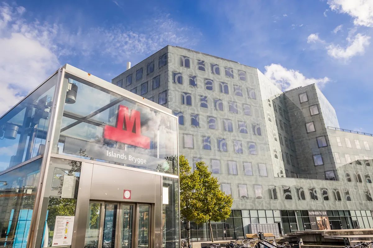 Store glassruder og rå stil med Islands Brygge metrostation i baggrunden, en facade med mange vinduer og moderne design samt parkerede cykler foran bygningen