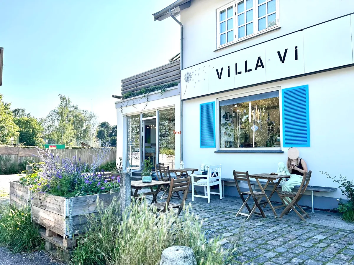 På Kongevejen 61 i Kongens Lyngby ligger en hyggelig café med have. En kvinde nyder en morgenmad på terrassen med blomster og træborde. Caféen har lysevinduer og blå rudesalter.