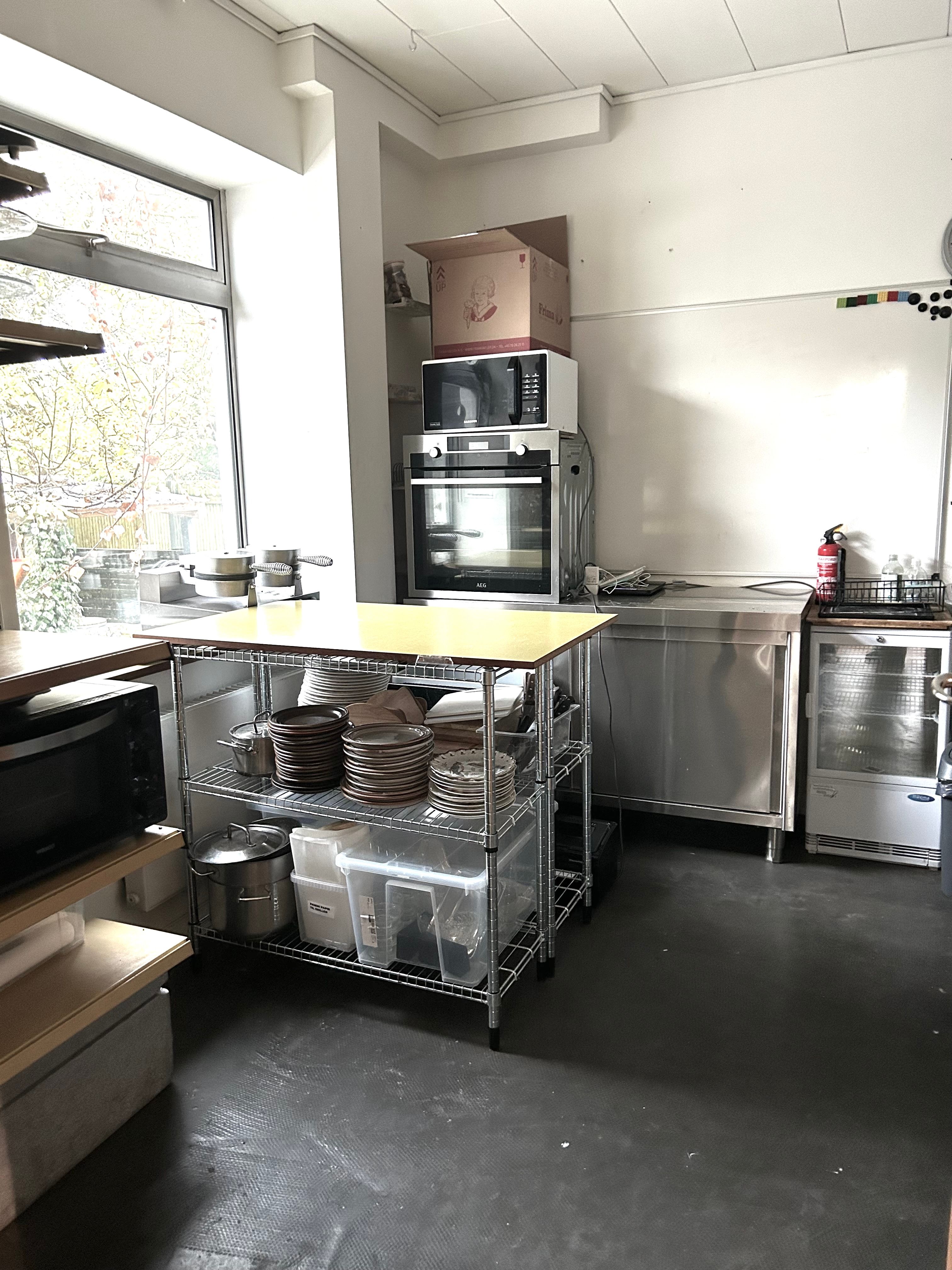 Køkken med to mikrobølgeovne, komfur, ovn og køleskab, træbord med tallerkener, gryder og kugleglas samt en række rustfri stålreoler og et ildslukker