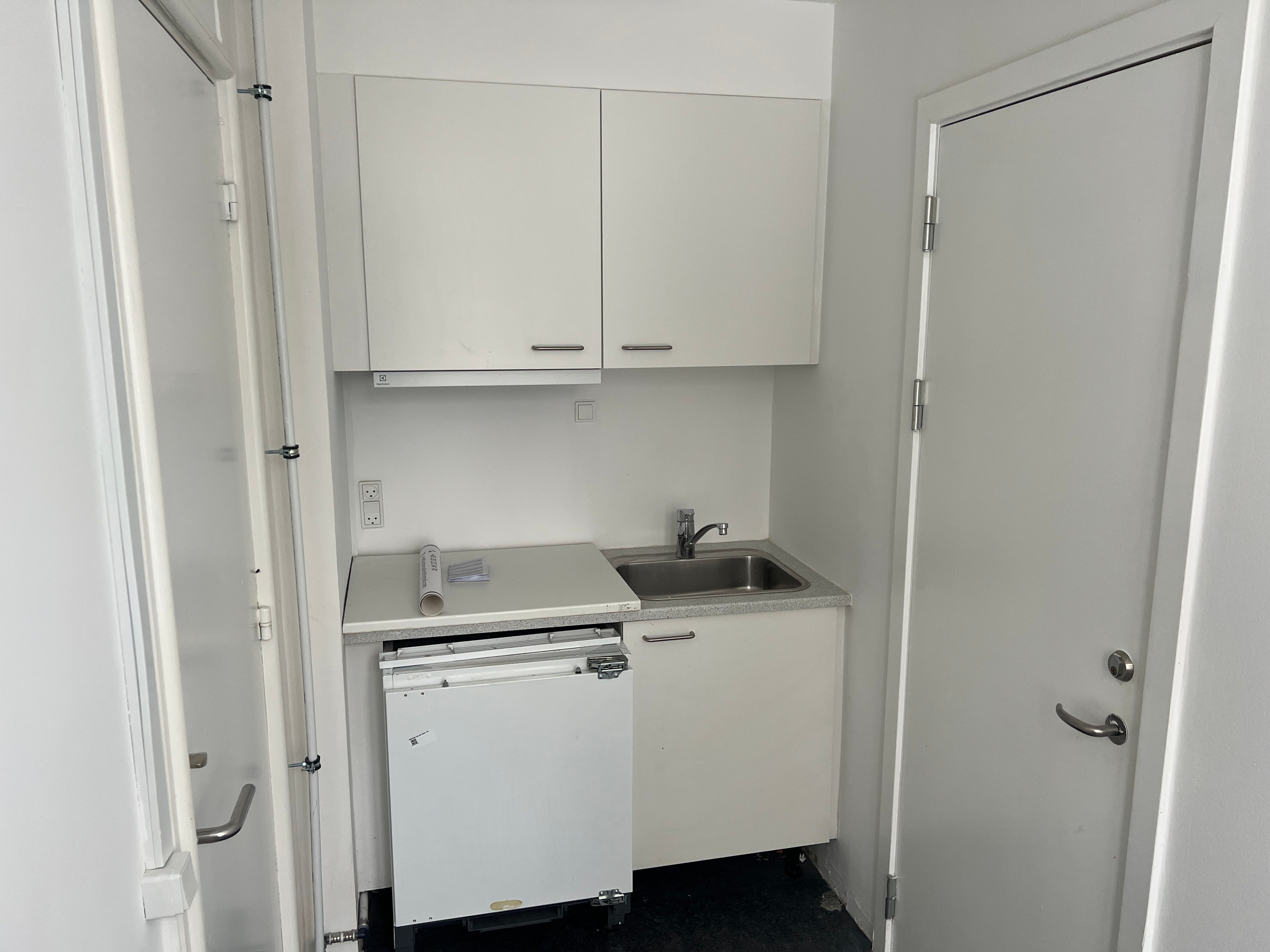 Foto af et lille køkken med hvide vægge og skabe, en enkel vask og en hvid ovn i hjørnet