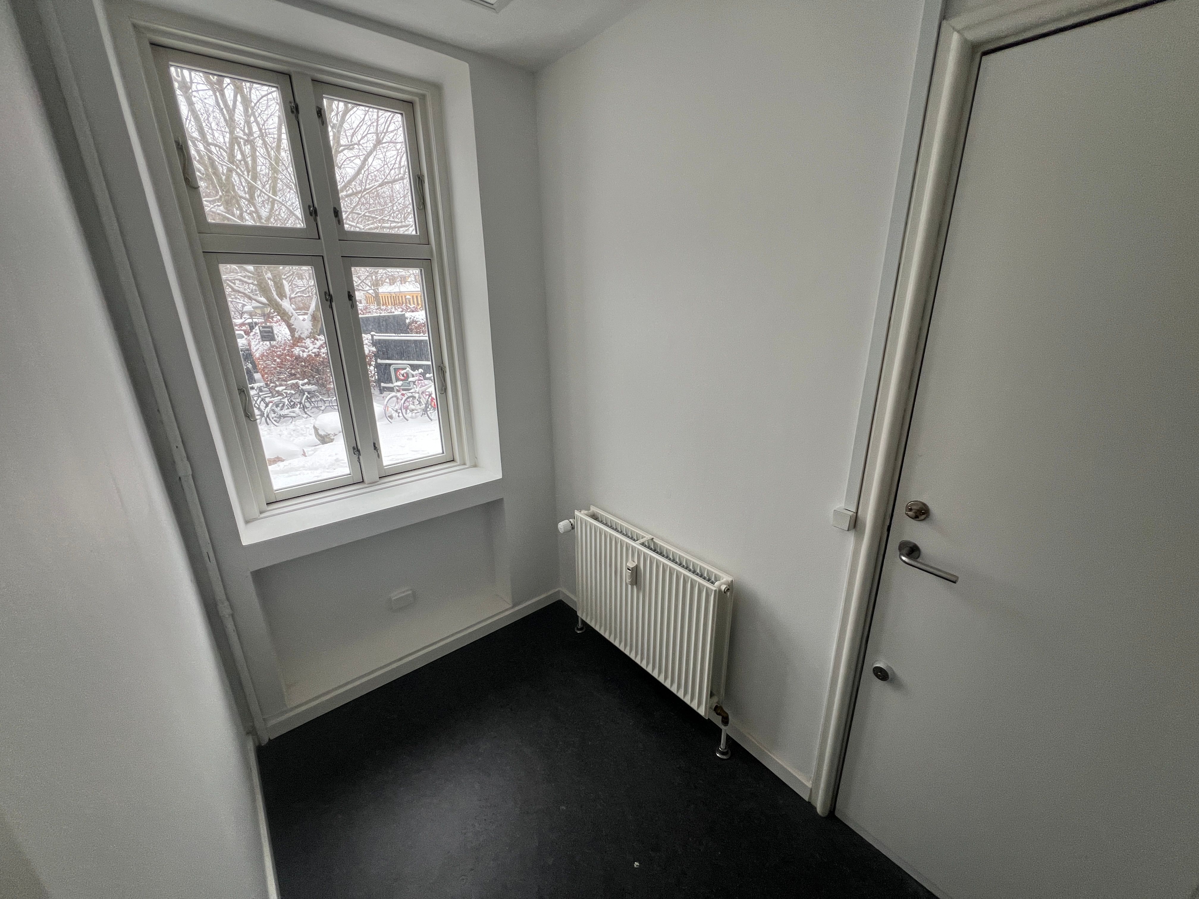 Et lyse og moderne lejemål med hvide vægge og en stor vinduesfront i hjørnet af lokalet. En radiator er installeret på væggen, og der er sne udenfor.