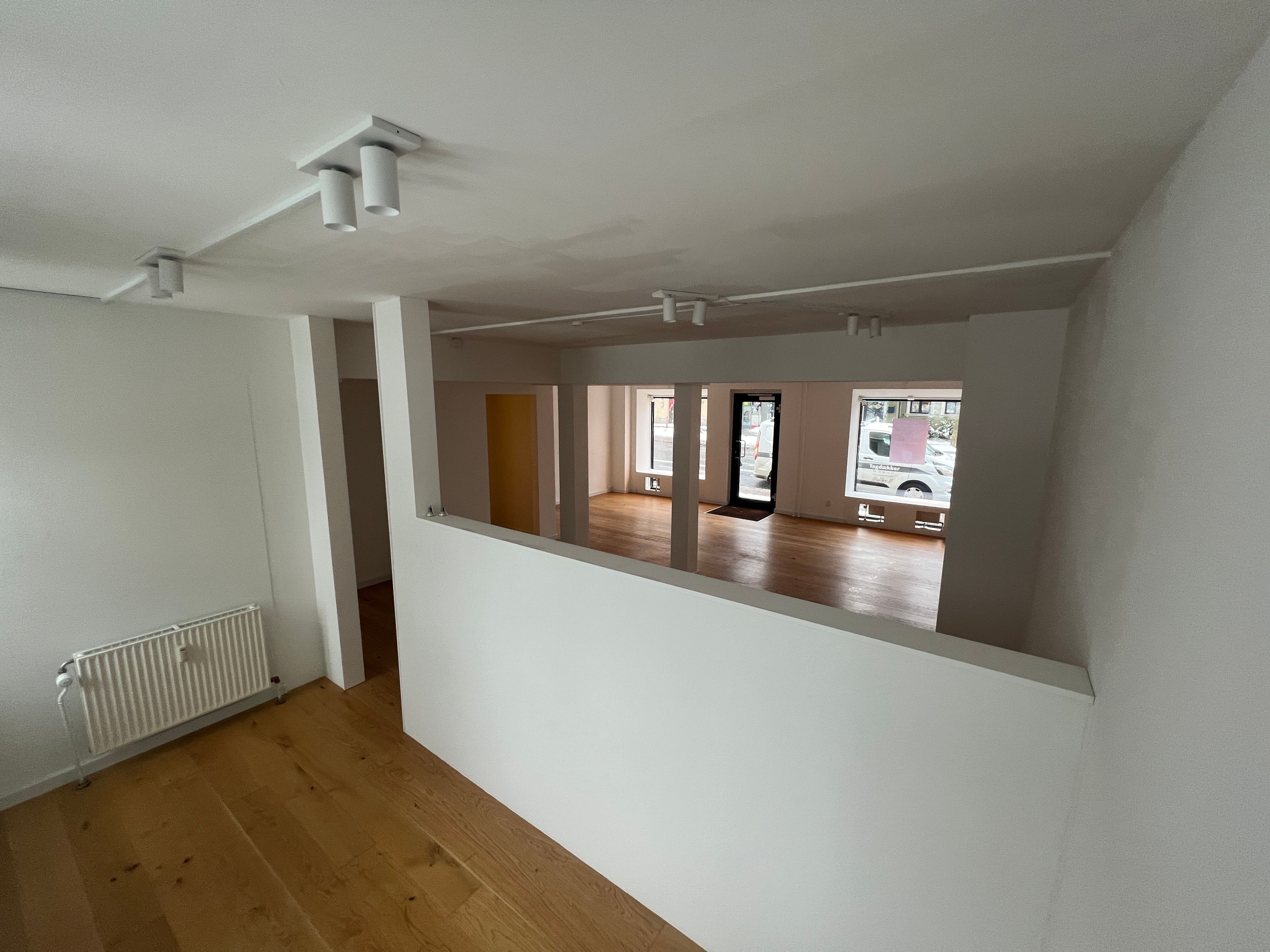 Indeni et tomt lokale med hvide vægge og lysegule trægulve. Et hvidt radiator er placeret på den ene side af lokalet. Foroven er der et lille vindue med en pink tekst.