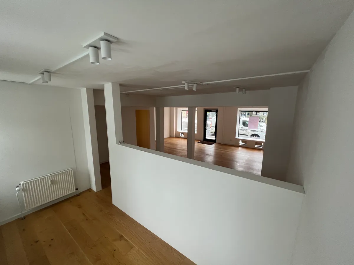 Indeni et tomt lokale med hvide vægge og lysegule trægulve. Et hvidt radiator er placeret på den ene side af lokalet. Foroven er der et lille vindue med en pink tekst.