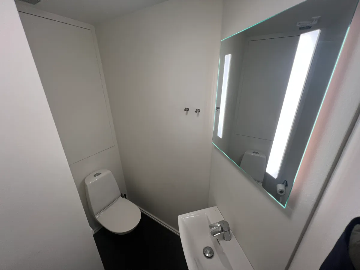 Et lille, hvidt badeværelse med toiletter, hvidt toilet og en stor spejl med indbygget lys.