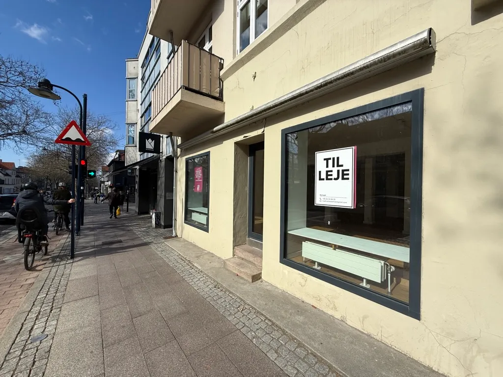 Mennesker kører cykler og går på gaden, hvor der er en schild for "Til leje" i butiksvinduet og rødt stoplys