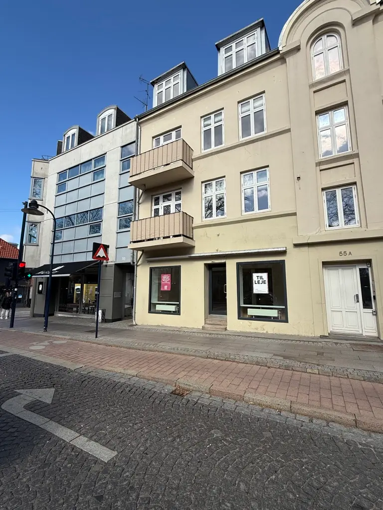 En butik til leje på Lyngby Hovedgade 55A med hvide vægge, røde mursten og tre balkoner. Adgang til gaden med hvidt gulv og en vejvisningsskilt