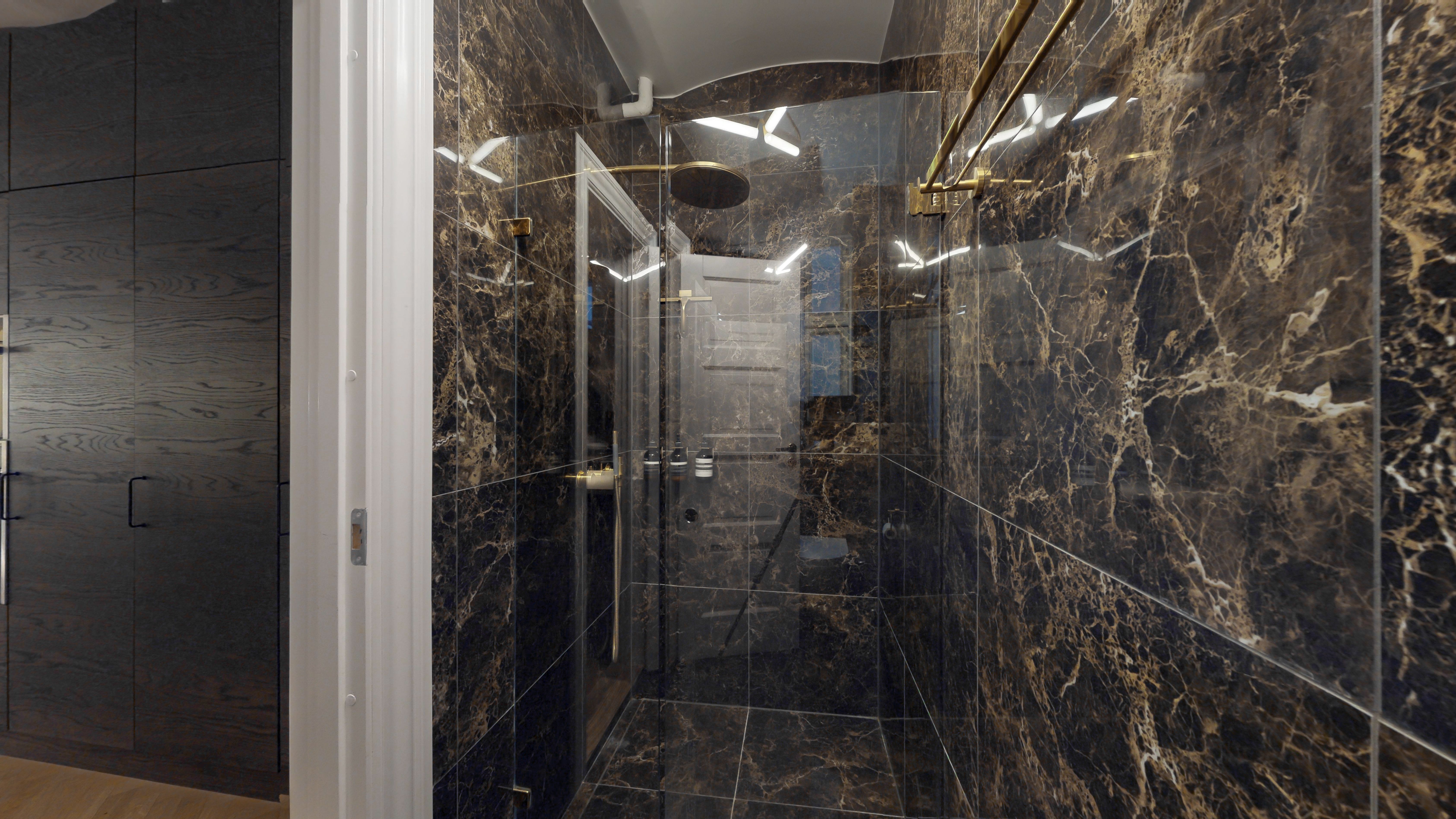 Foto af et moderne badeværelse med marmorbeklædning, guldfarvede håndtaget, og en glashåndbruser med indbygget ventil. Lyse hjørner og elegante designelementer giver rummet et luksuriøst udseende