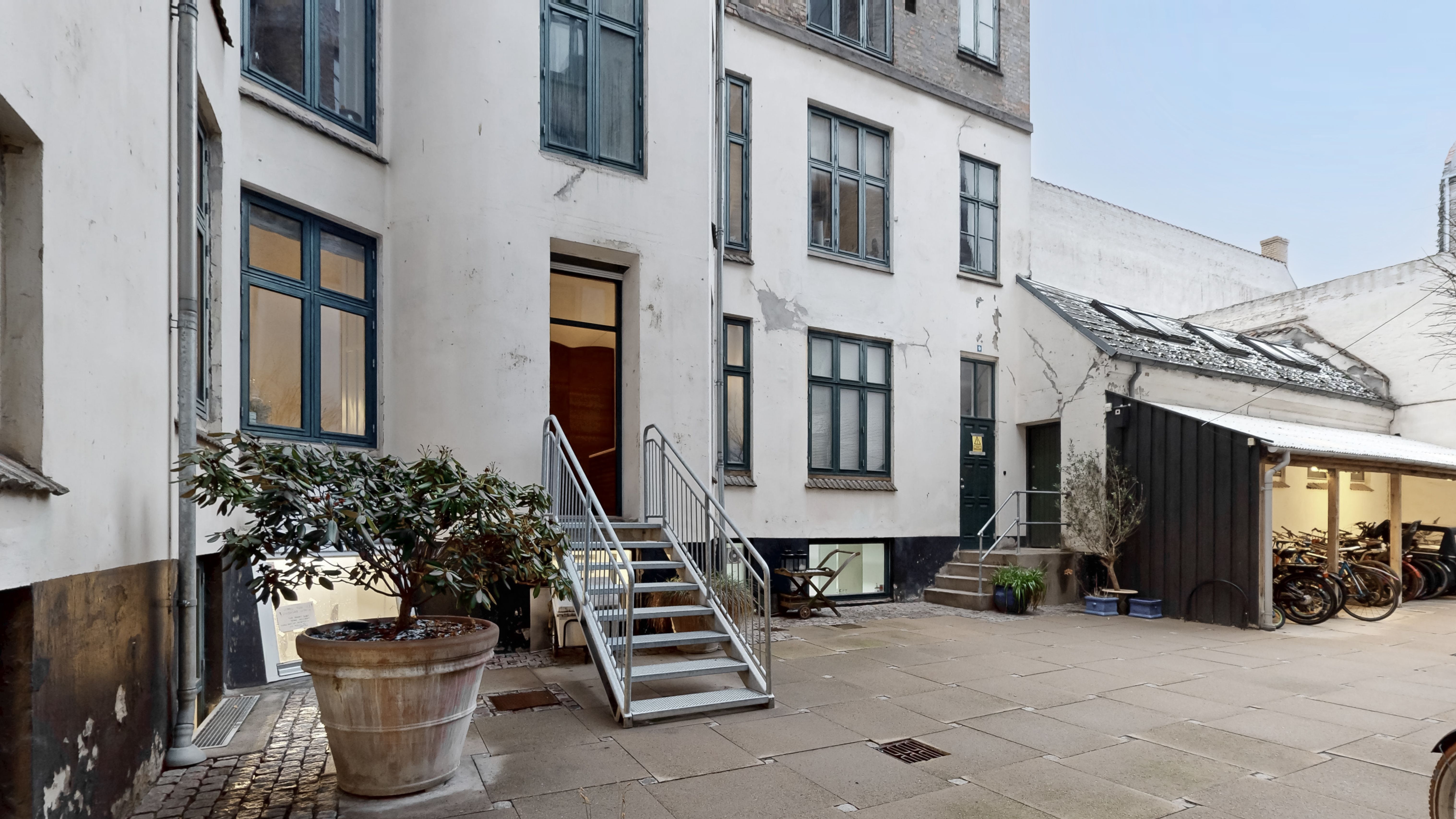 Erhvervsandel med store ruder og terrasse, der vender ud mod en fredelig gade med parkerede cykler og grønne plante potter