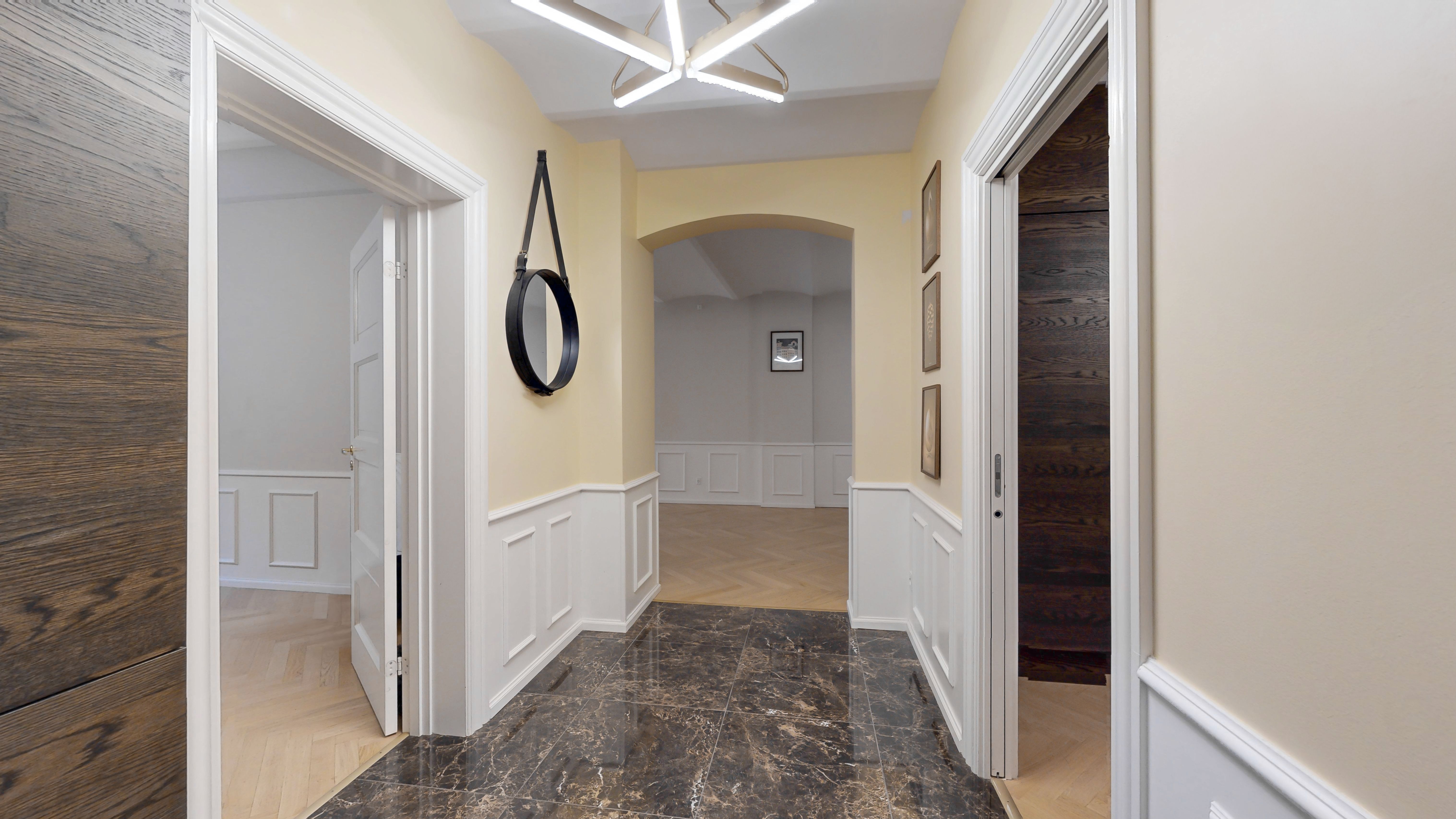 Indvendigt billede af en møbleret korridor med marmorgulv, hvide paneler, to spejle og tre rammer på væggen samt lyst gulv og lyse lofter