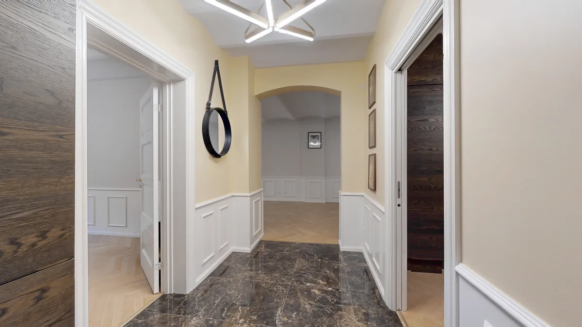 Indvendigt billede af en møbleret korridor med marmorgulv, hvide paneler, to spejle og tre rammer på væggen samt lyst gulv og lyse lofter