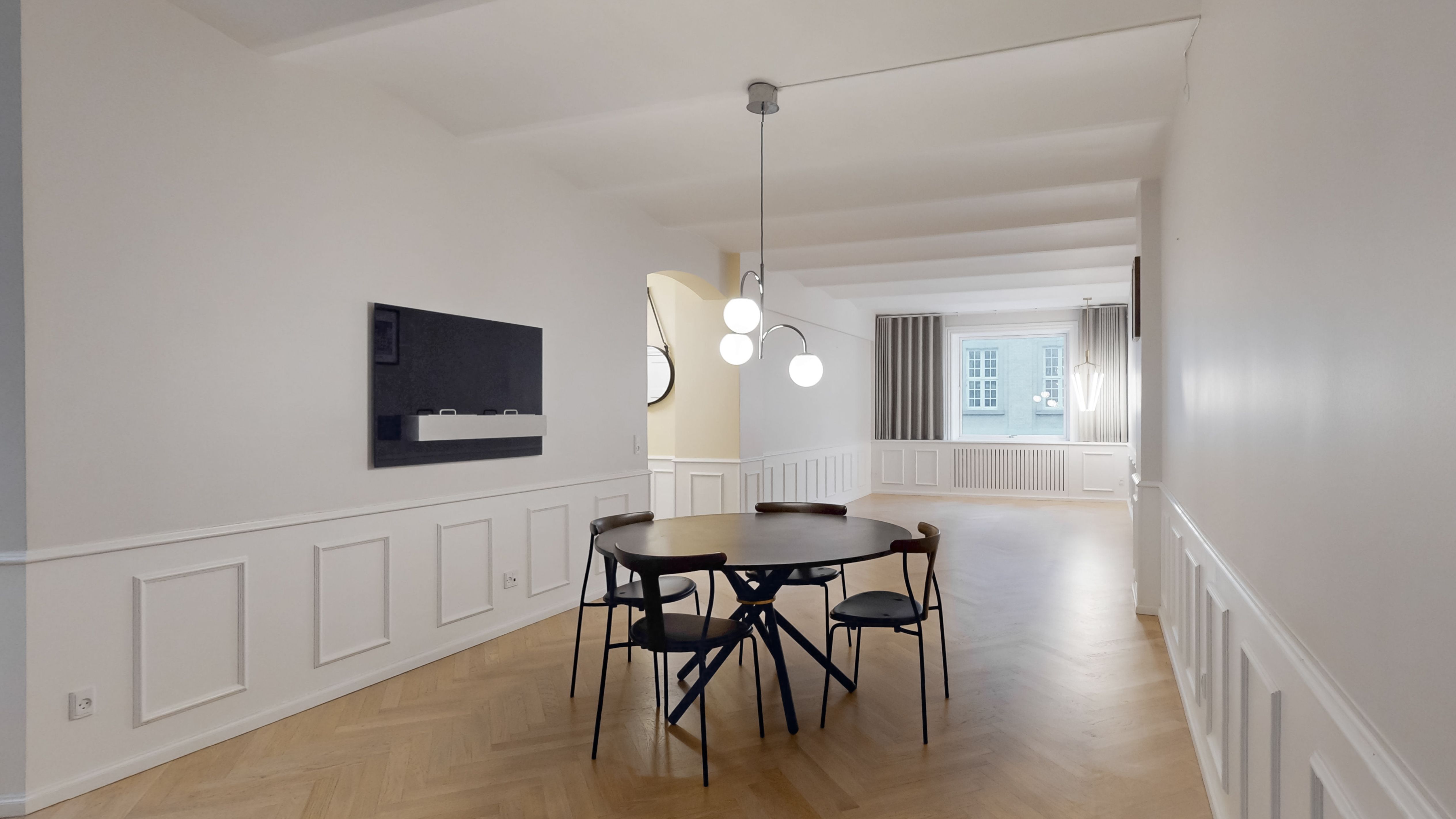 Minimalistisk stue med trægulv og rundt spisebord med fire stole, moderne lysekroner og store vinduer med gardiner samt højt til loftet