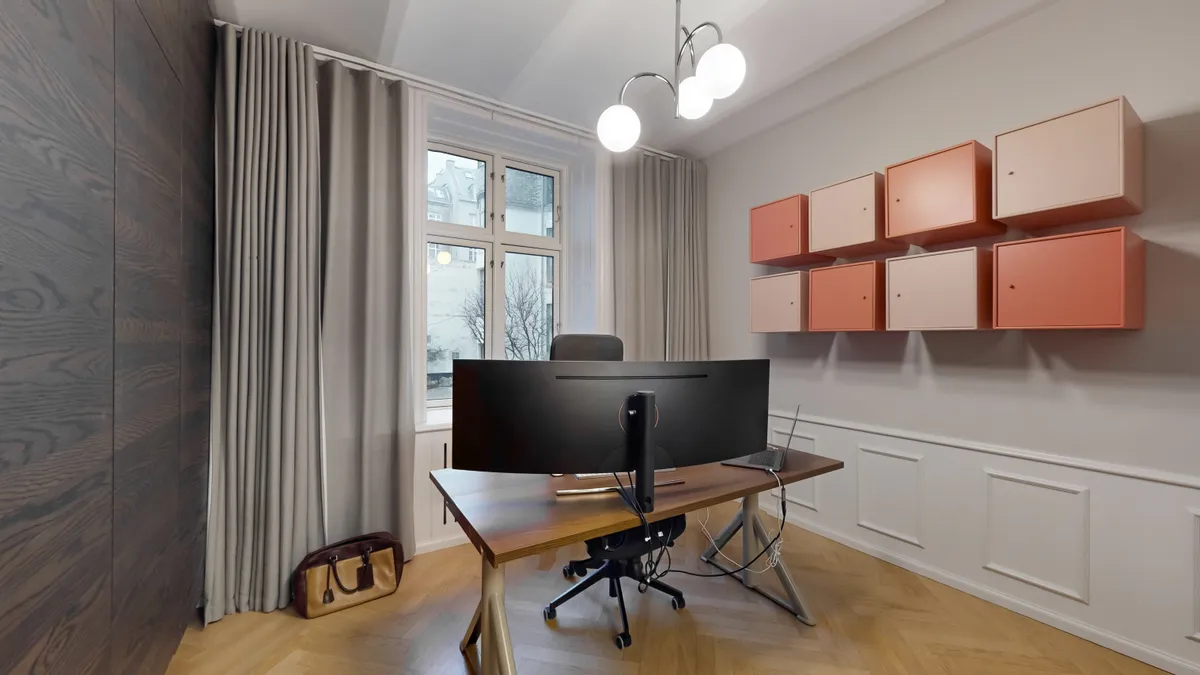 Et moderne kontorlokal med to stolpeformede lamper, en stor skærm og en bærbar computer samt en taske og en stol