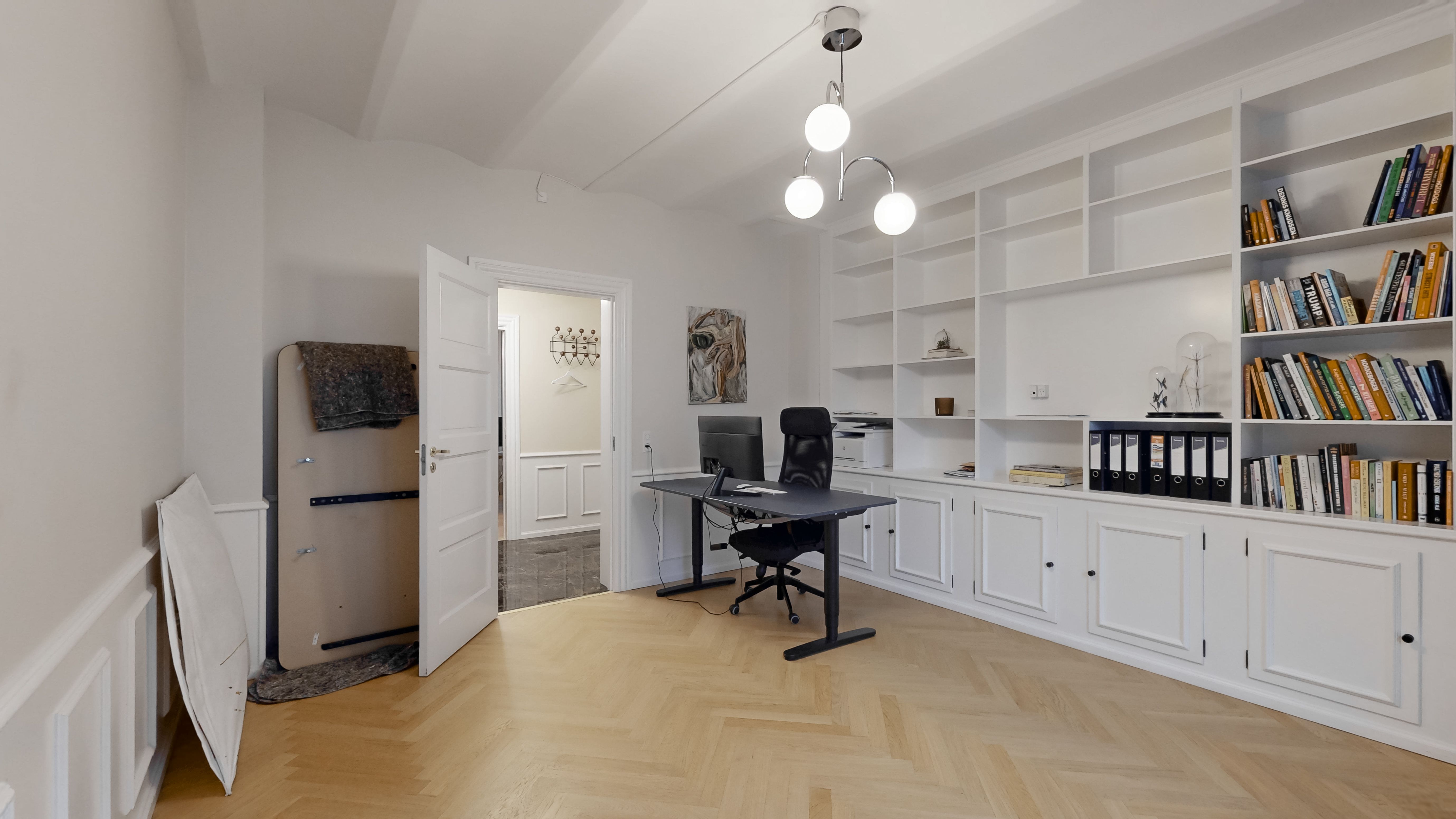 Kontor med lyse vægge, trægulv og høje hylder med bøger, møbler og en skrivebord med computer og stol.