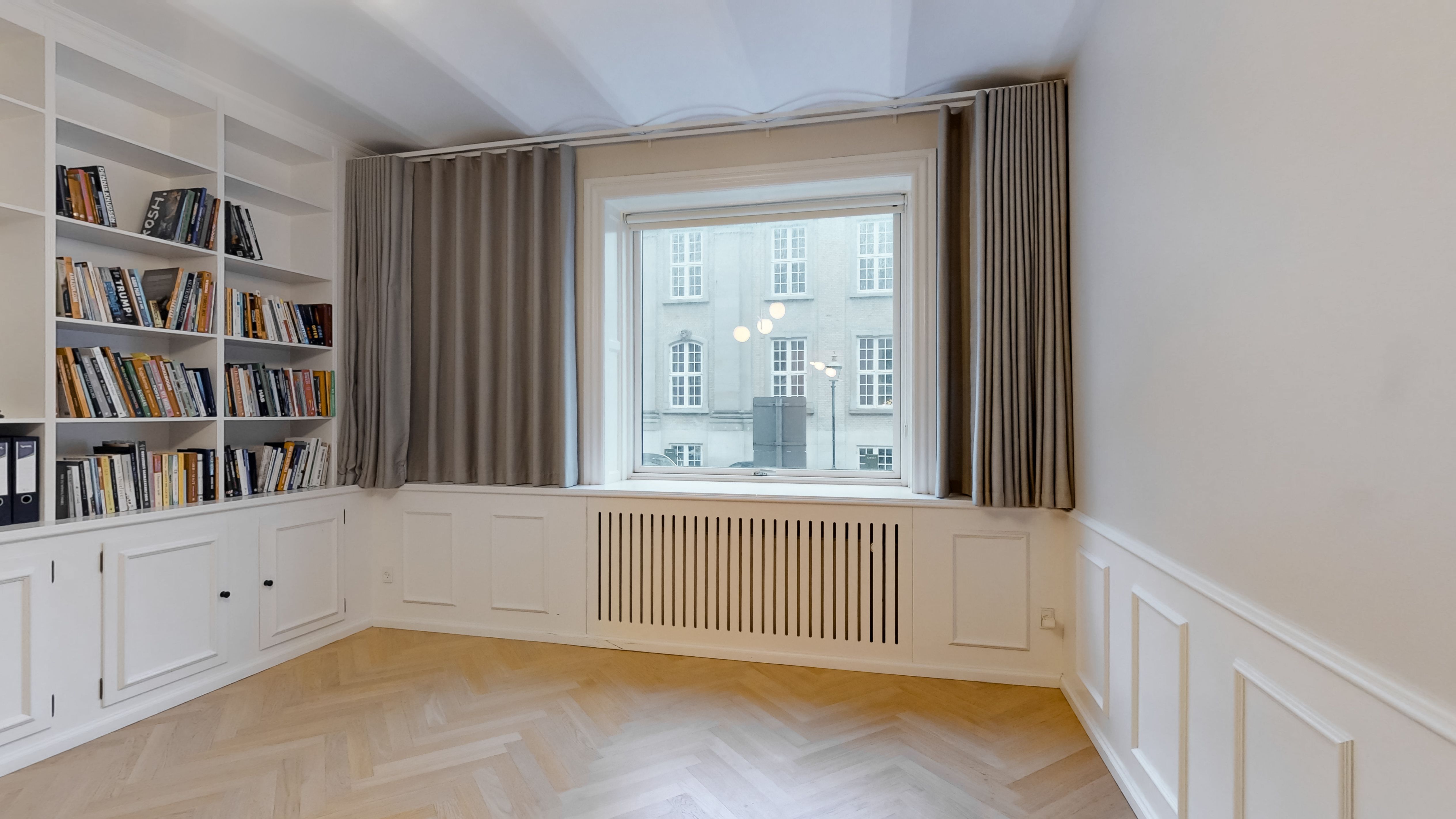 Hjørneværelse med lysegardiner og buevindue, hvide vægge og parketgulv samt højloft og indbygget radiator under vinduet