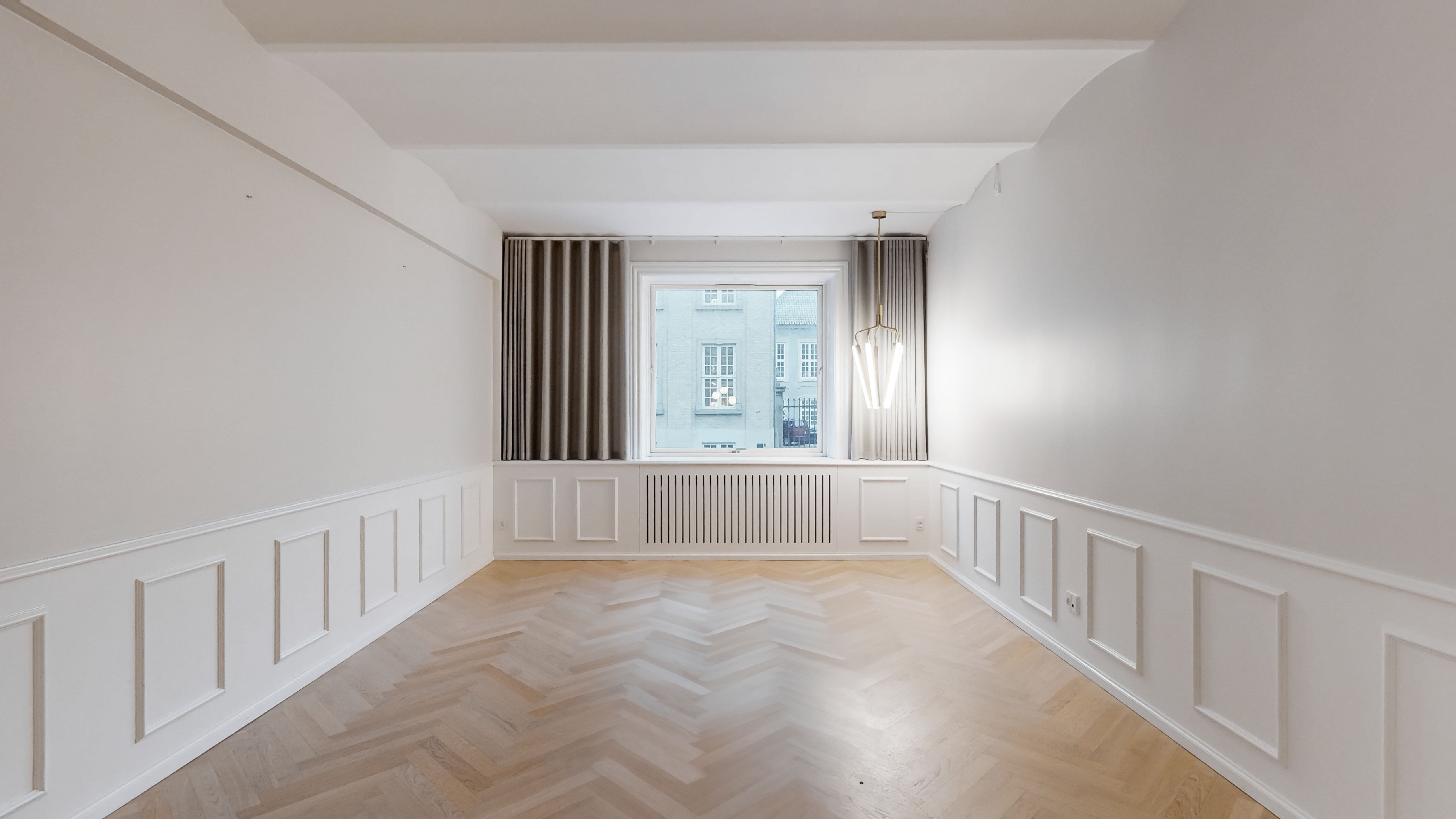 En tom rum med lyse vægge og trægulv. Et vindue med gardiner og et lysekroner i loftet. Væggene er dekoreret med paneler og der er en radiator under vinduet.