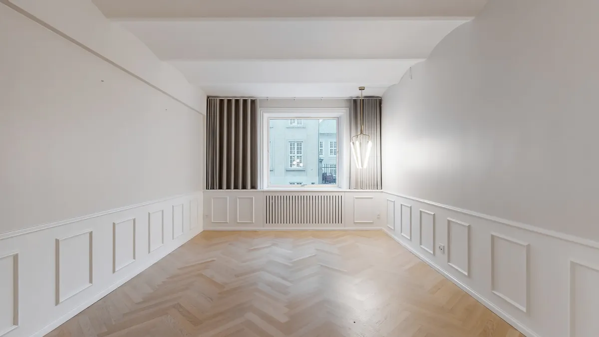 En tom rum med lyse vægge og trægulv. Et vindue med gardiner og et lysekroner i loftet. Væggene er dekoreret med paneler og der er en radiator under vinduet.