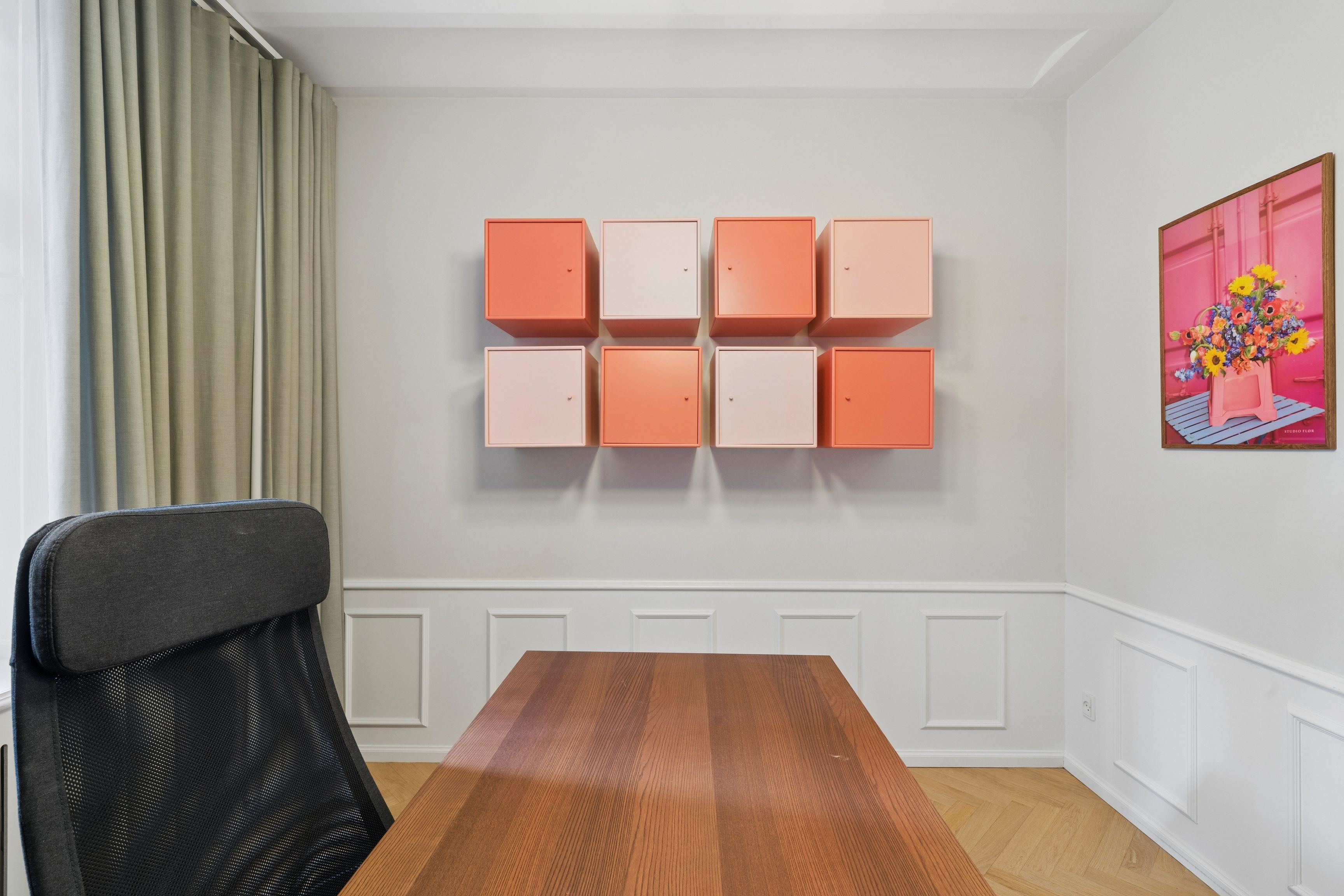 Værelse med orange og hvide loftsskabe og en lyse pink vægmaleri med en vase med blomster. Bord med en stol og en gardin i baggrunden