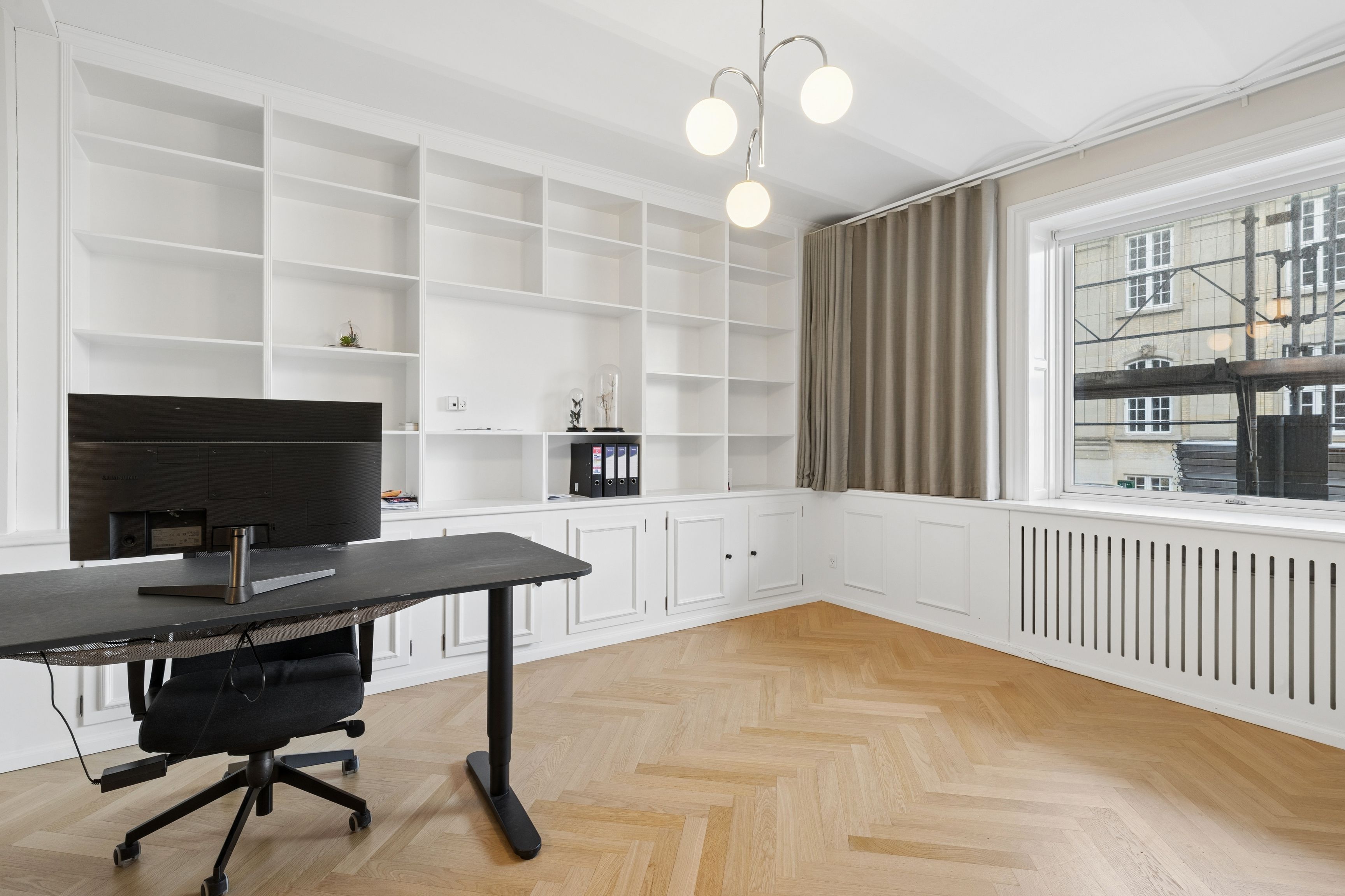 Studieværelse med højt loft, høj stue, moderne skrivebord med Samsung-skærm og højtalere, hvide vægge og hylder, trægulv, vinduesgardiner og udsigter til bygninger og elektriske ledninger