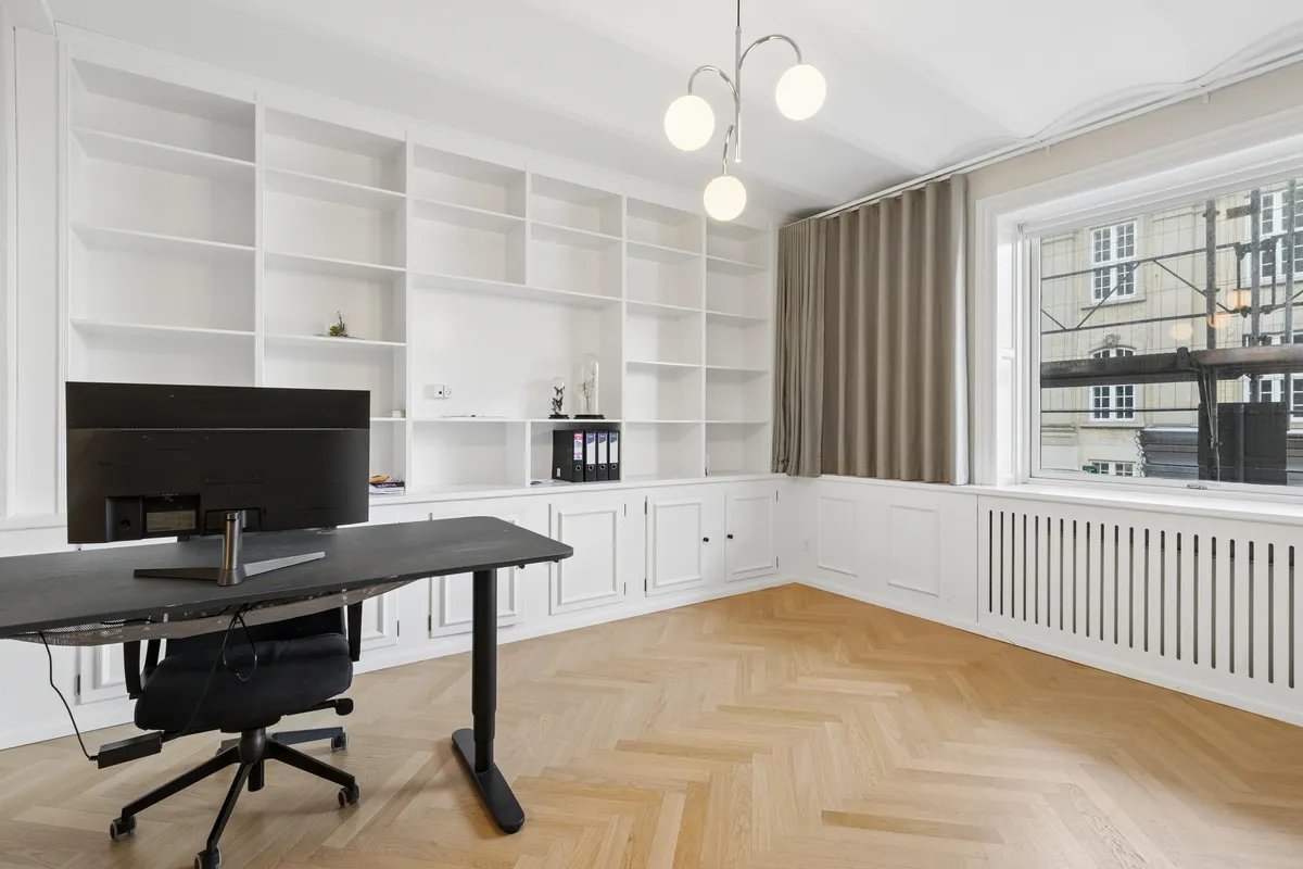 Studieværelse med højt loft, høj stue, moderne skrivebord med Samsung-skærm og højtalere, hvide vægge og hylder, trægulv, vinduesgardiner og udsigter til bygninger og elektriske ledninger