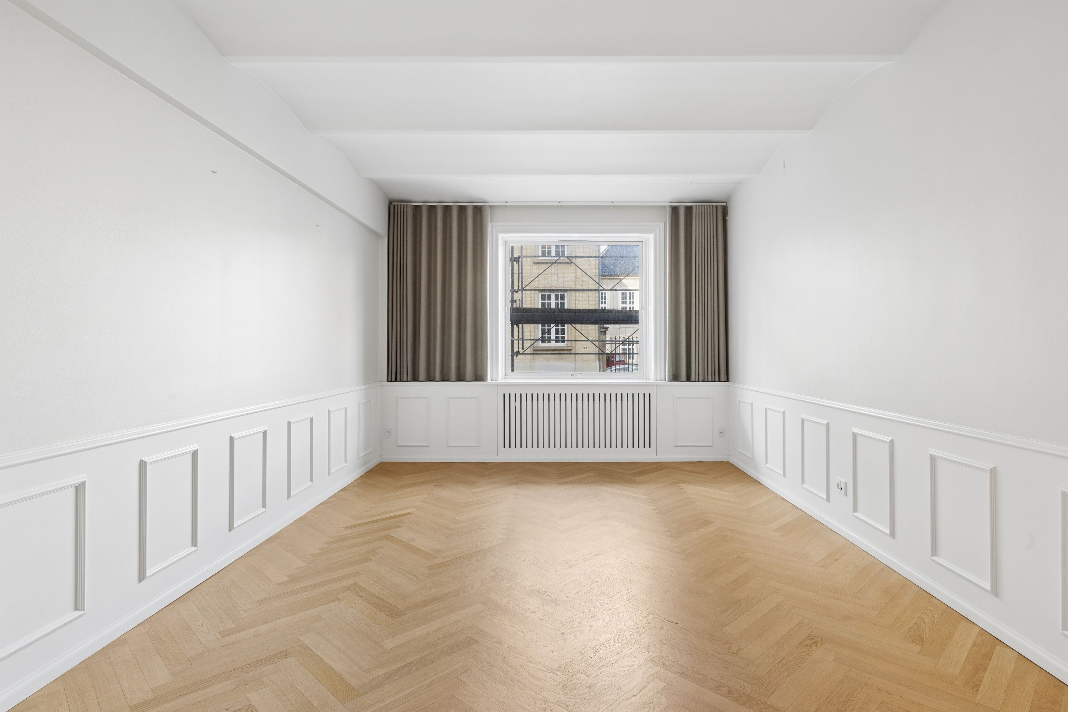 Hvidt værelse med lysegul trægulv og hvide vægge. Rummet er delt med en hvid skab og en række hvide paneler. Vindue med mørkegardiner.