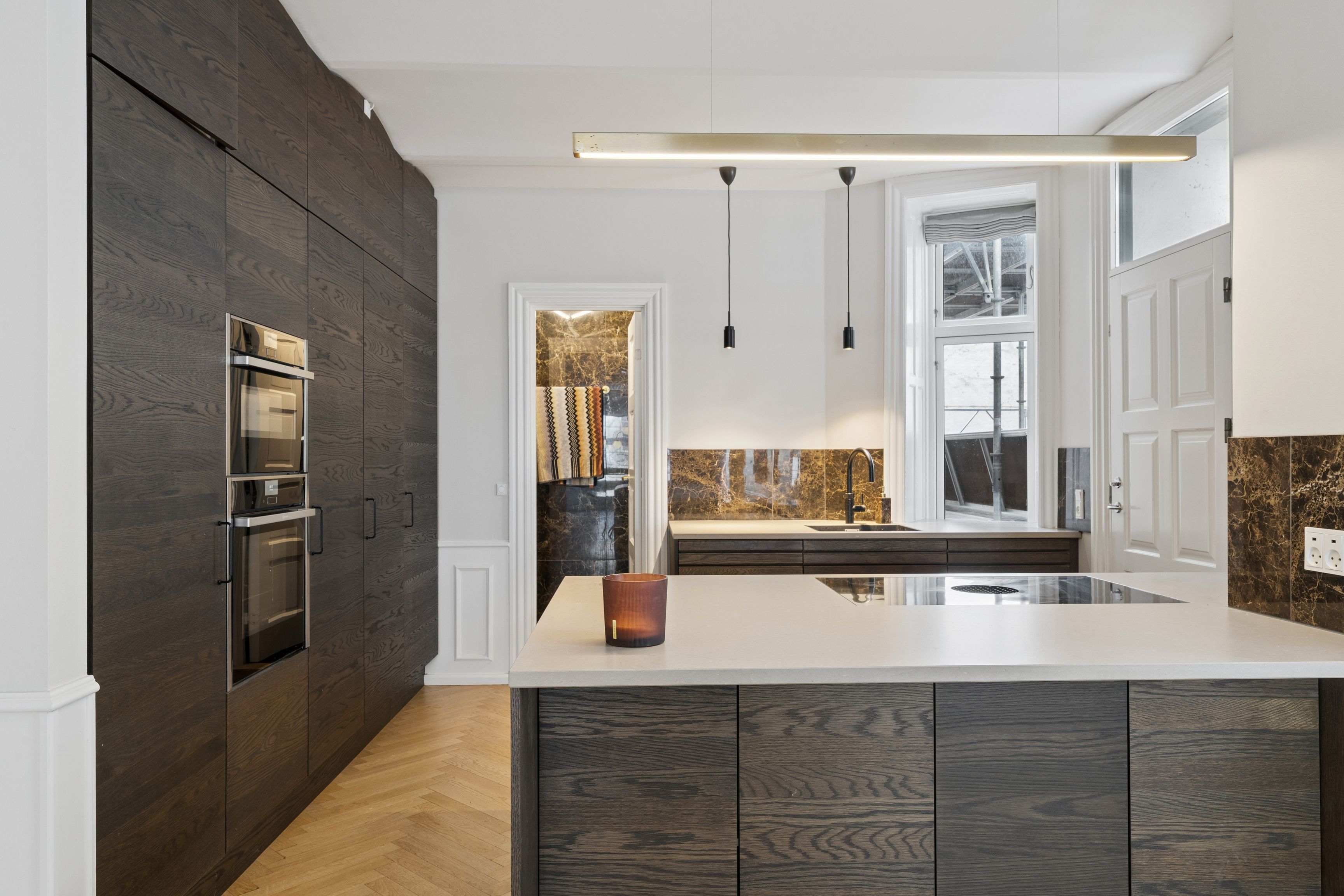 Moderne køkken med mørke træskabe og hvid marmor arbejdstavle og gulv, lys over ovnen, høj lysekron og åbne døre til marmor og møbelskabe