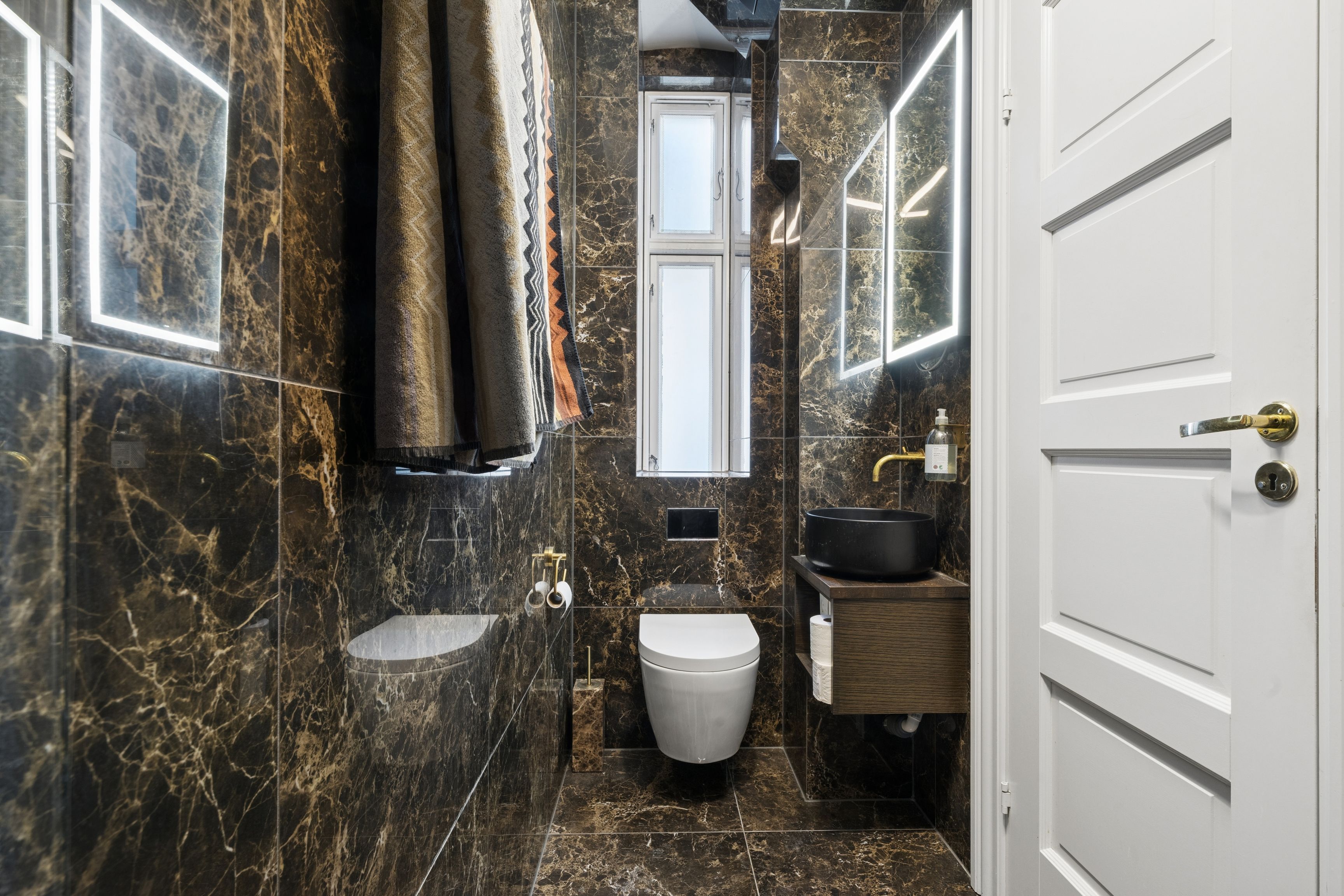 Et moderne badeværelse med marmorbelægning, hvidt toilet og vask samt en åben dør og vinduesruder.